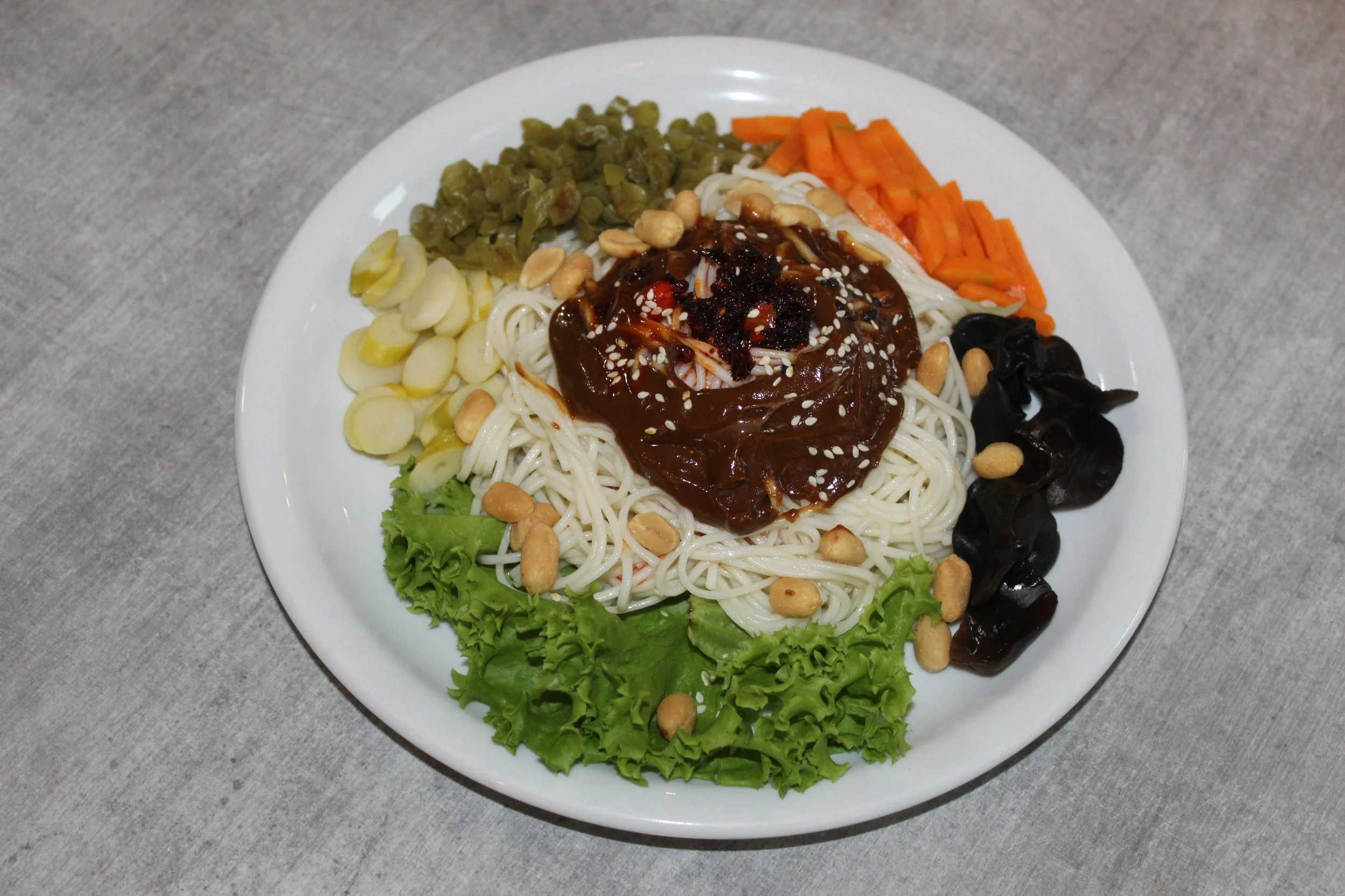 A plate of Korean cold noodles with various vegetables and toppings, including shredded carrots, black wood ear mushrooms, chopped garlic, pickled radish, chopped green beans, lettuce, and a brown sauce with sesame seeds and crushed peanuts on top.