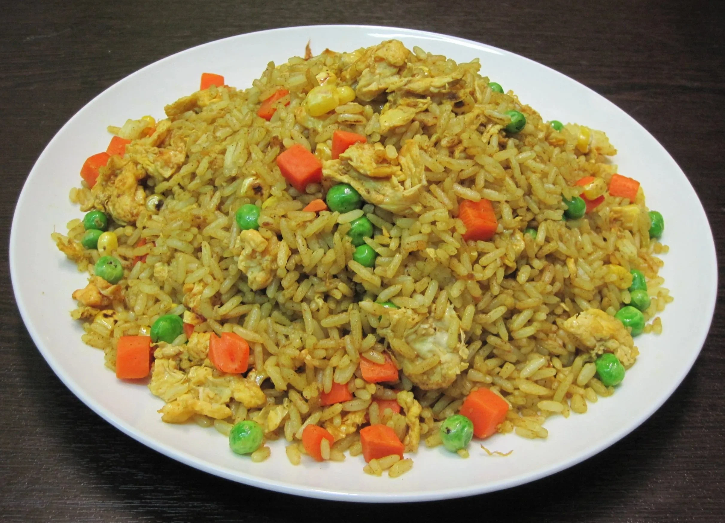 A plate of fried rice with mixed vegetables, including peas and diced carrots, garnished on a dark wooden surface.