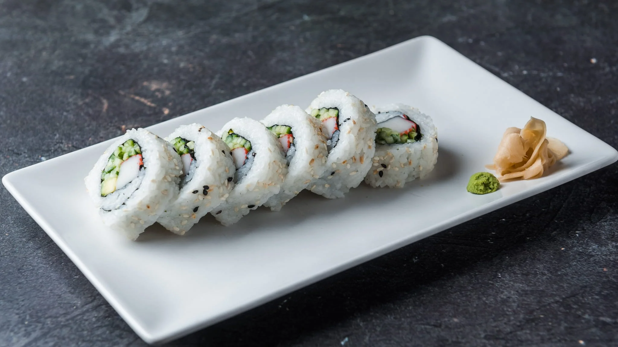 Sushi rolls with avocado, cucumber, and imitation crab on a white rectangular plate, garnished with pickled ginger and wasabi, on a dark textured surface.