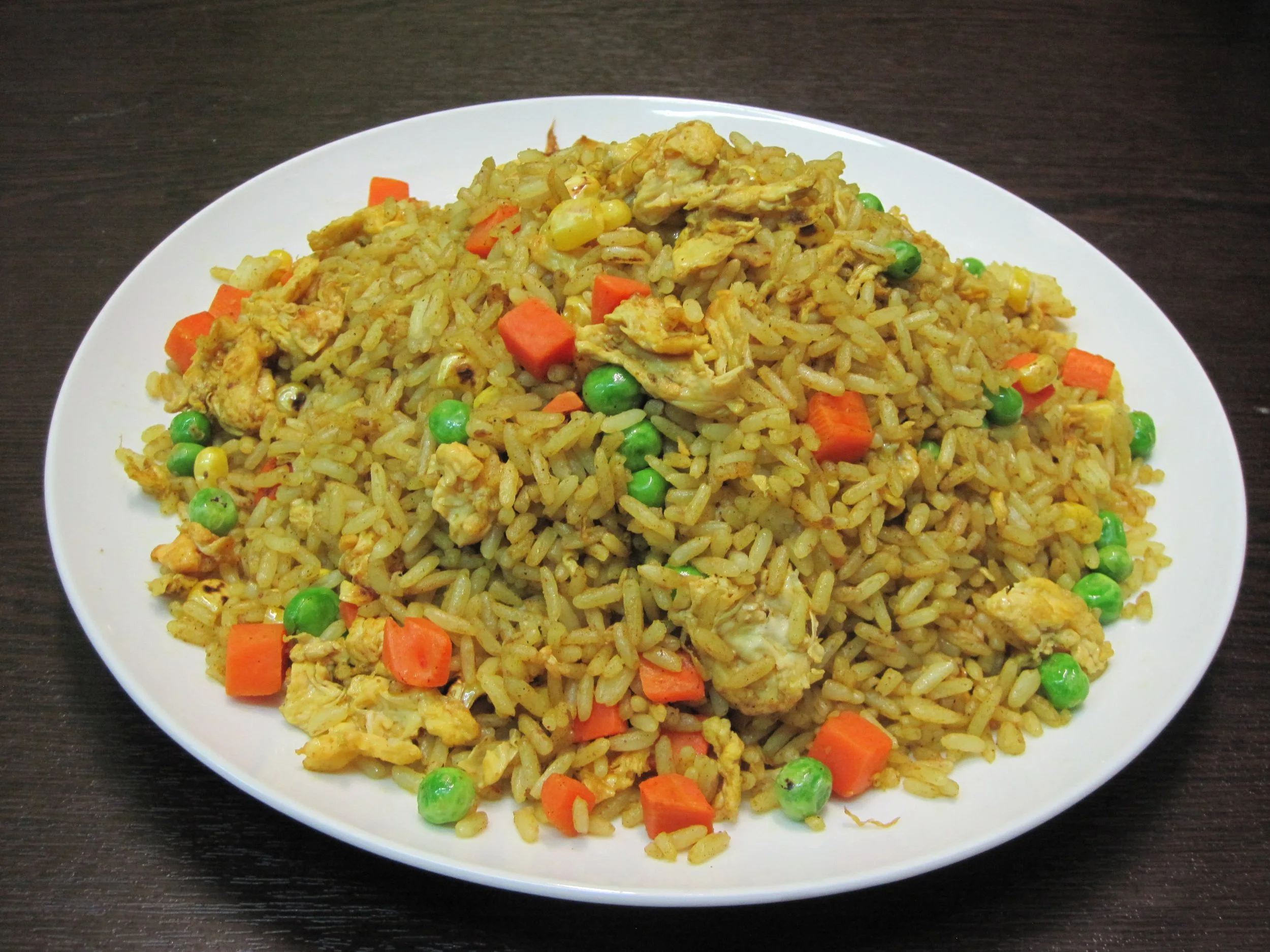 A white plate of fried rice with peas, carrots, and scrambled eggs on a dark wooden table.