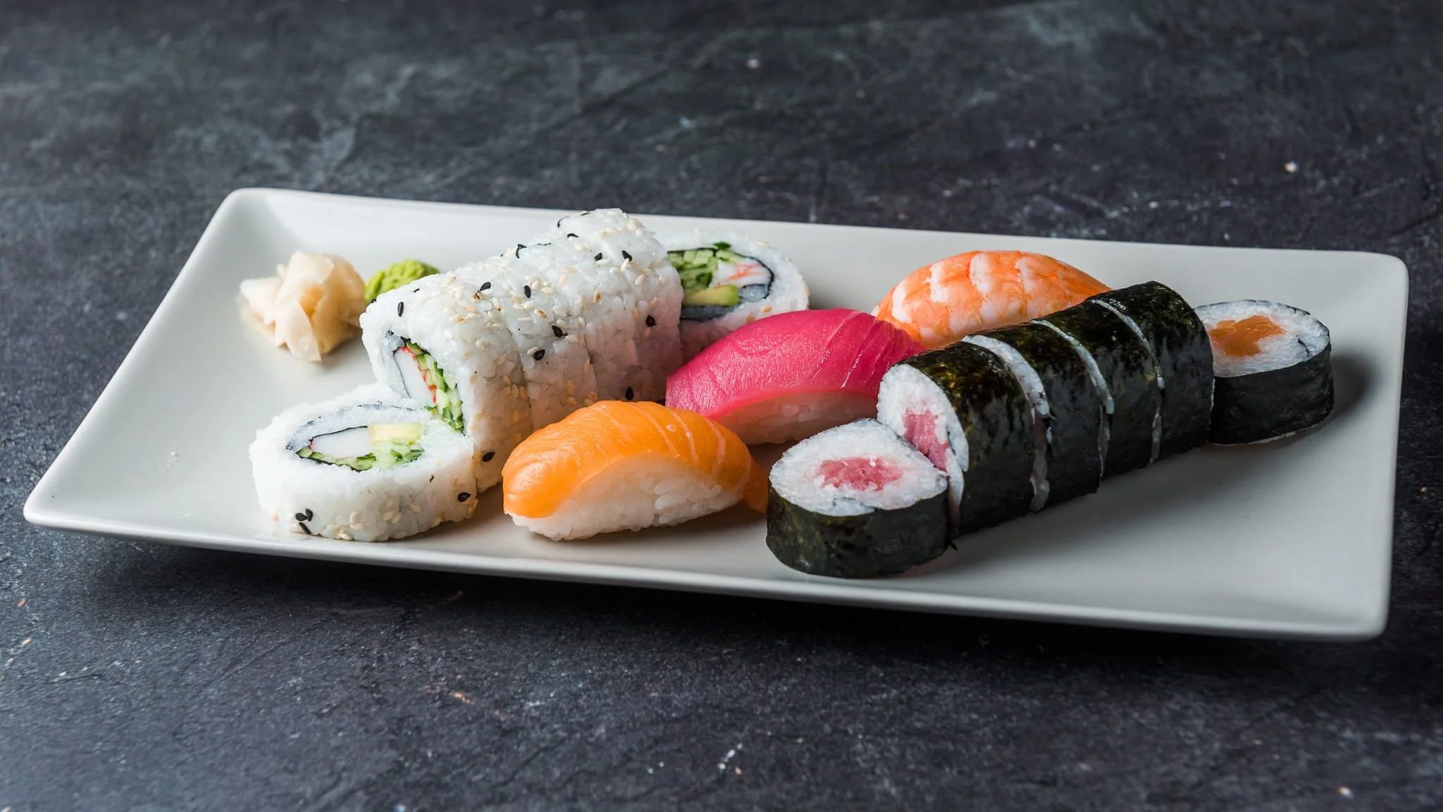 Assorted sushi rolls and sashimi on a rectangular white plate, with ginger and wasabi on the side, placed on a dark textured surface.