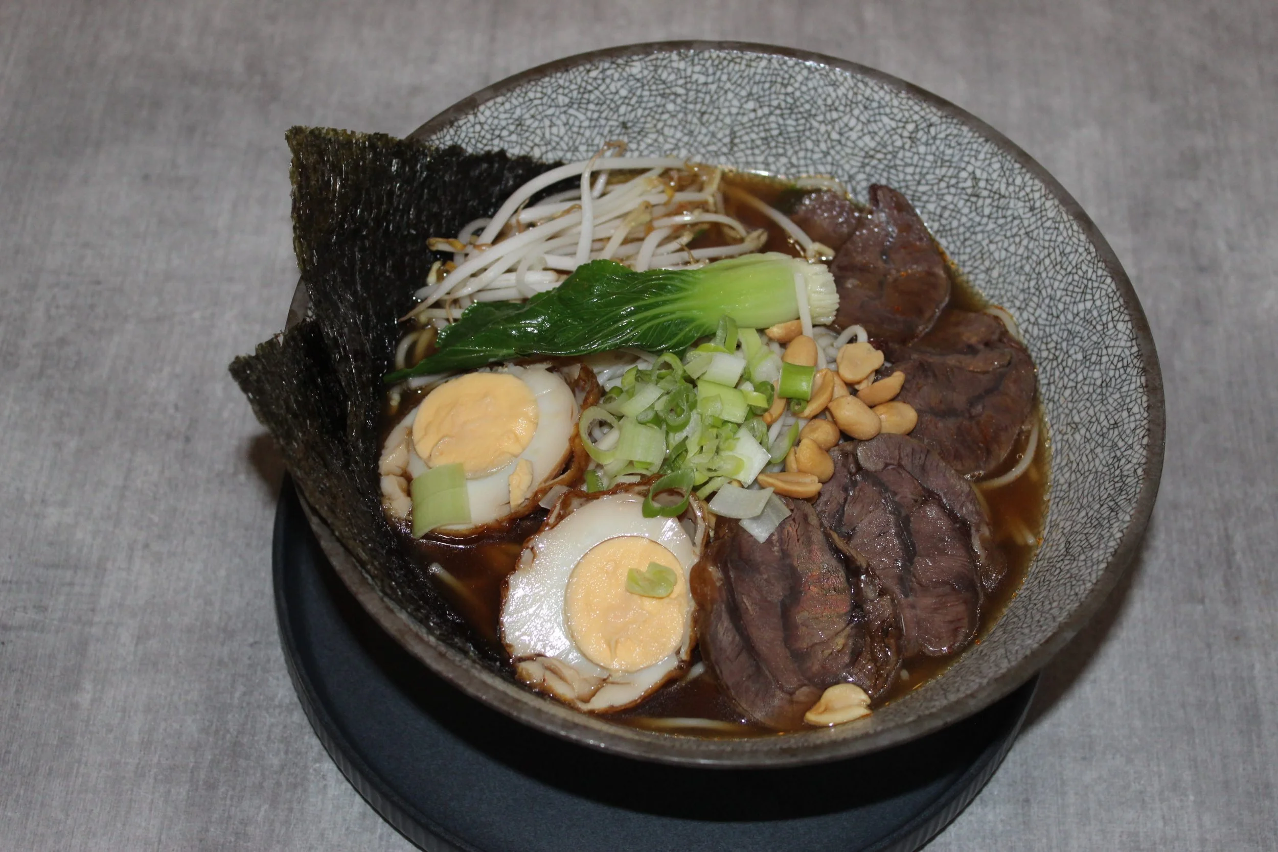Bowl of ramen with boiled eggs, beef slices, green onion, bean sprouts, seaweed, and peanuts.