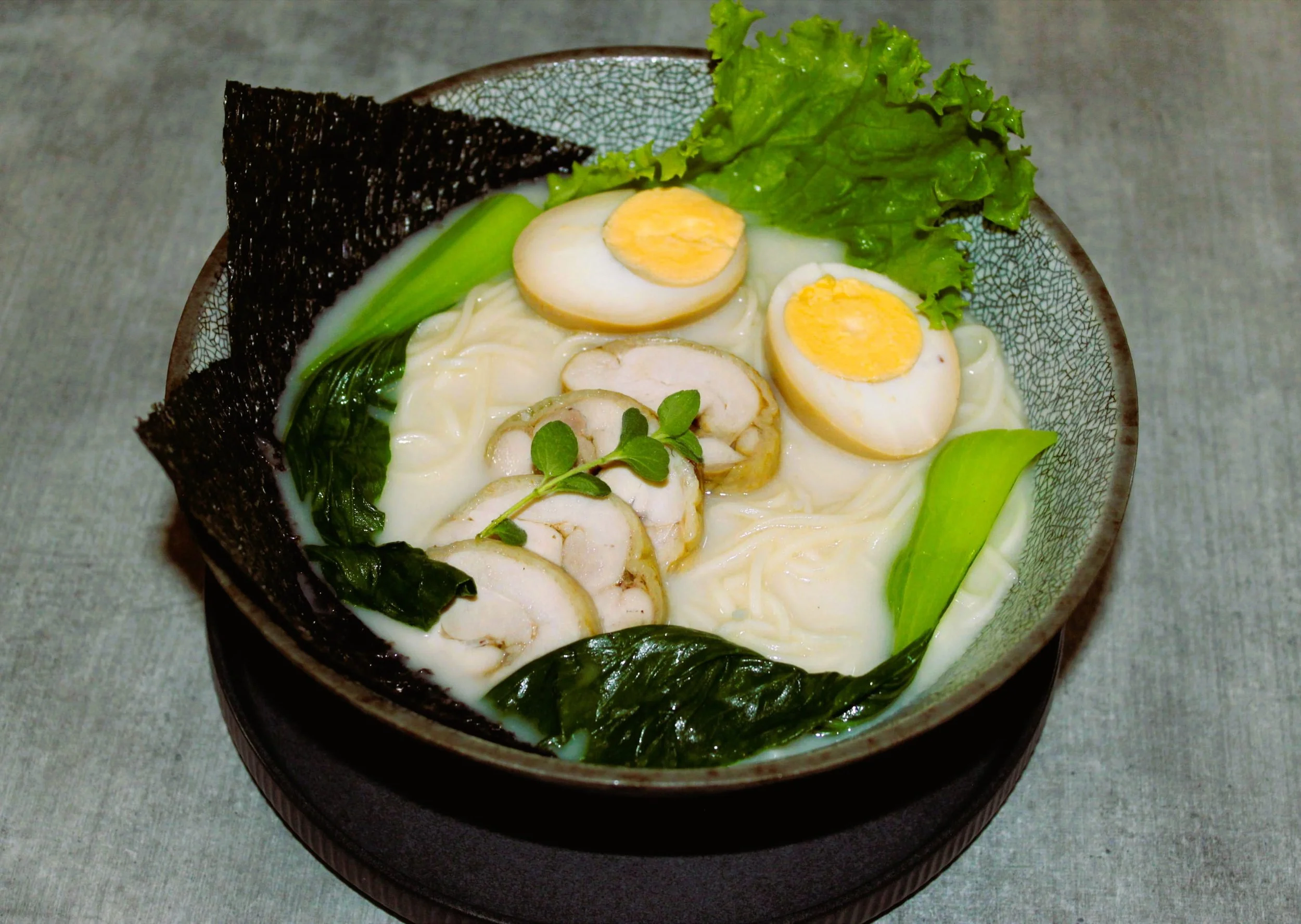 Bowl of ramen noodles with sliced chicken, soft-boiled eggs, leafy greens, and seaweed