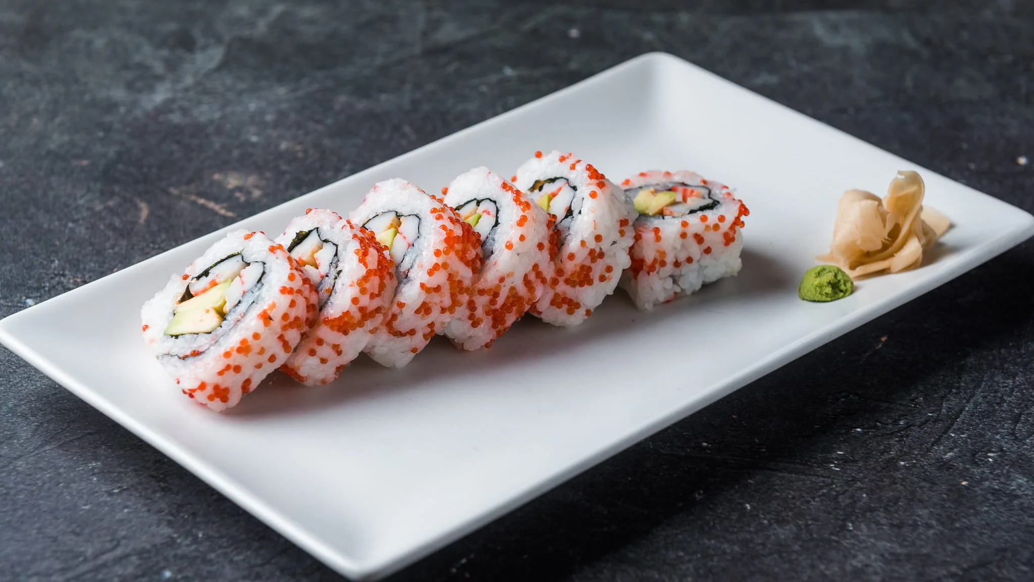 Sushi rolls with orange fish roe, avocado, and cucumber, served on a white rectangular plate with pickled ginger and wasabi, on a dark textured surface.
