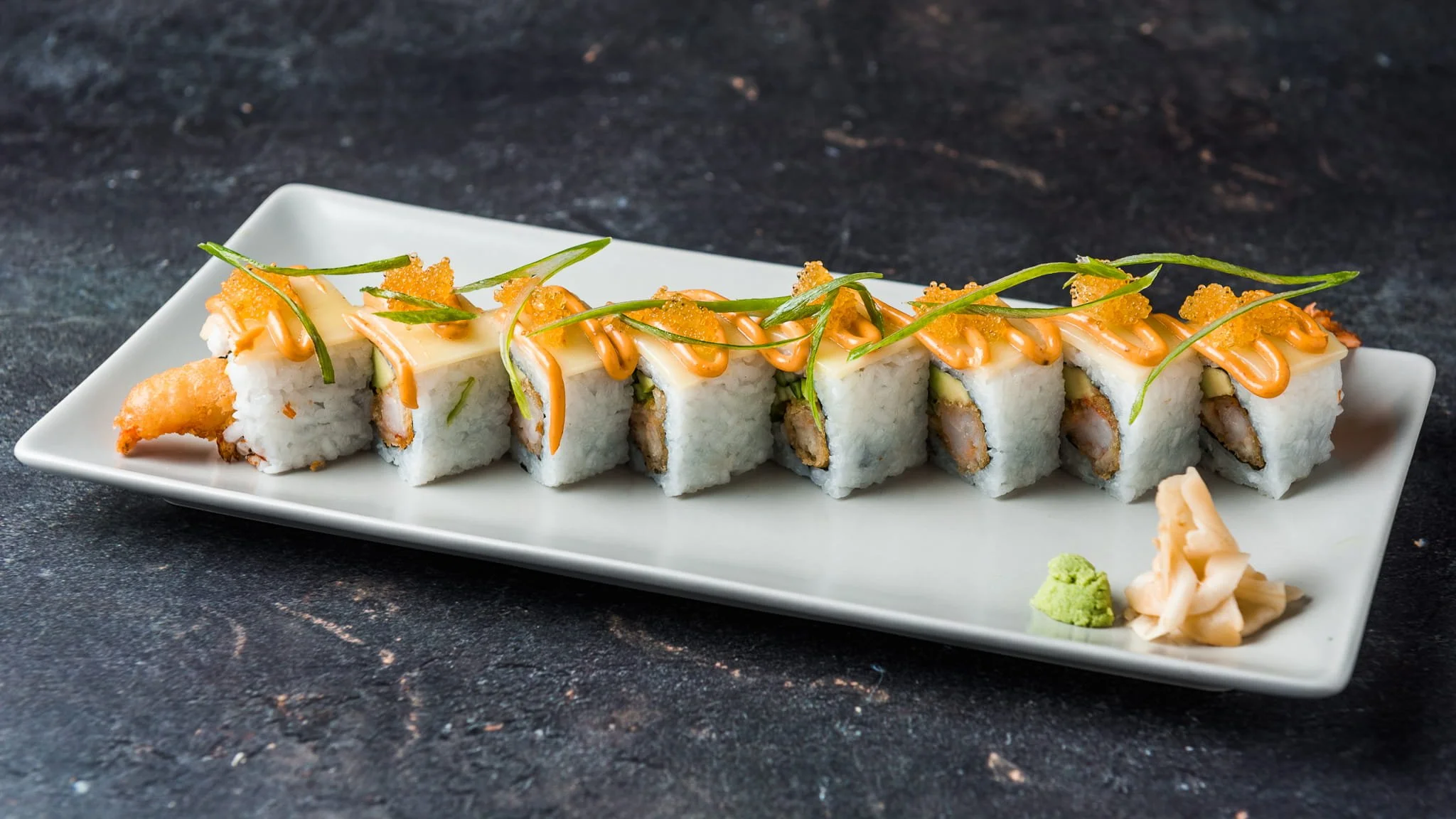 Sushi roll on a rectangular white plate, topped with orange sauce, thin green onion strips, and fish roe, served with wasabi and pickled ginger on the side. Dark textured background.