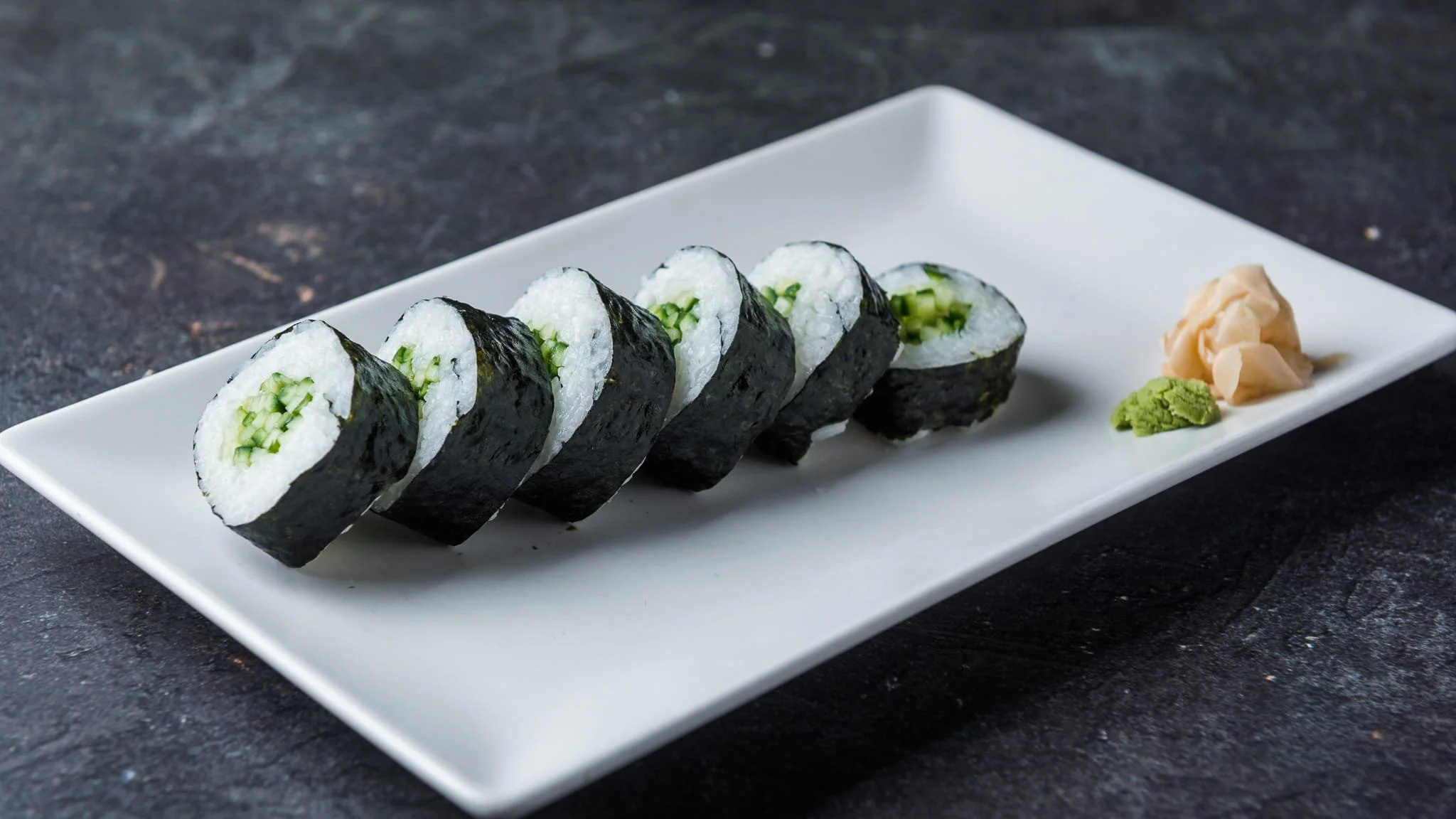 Six pieces of cucumber sushi rolls with nori seaweed wrapping, served with pickled ginger and wasabi on a white rectangular plate.