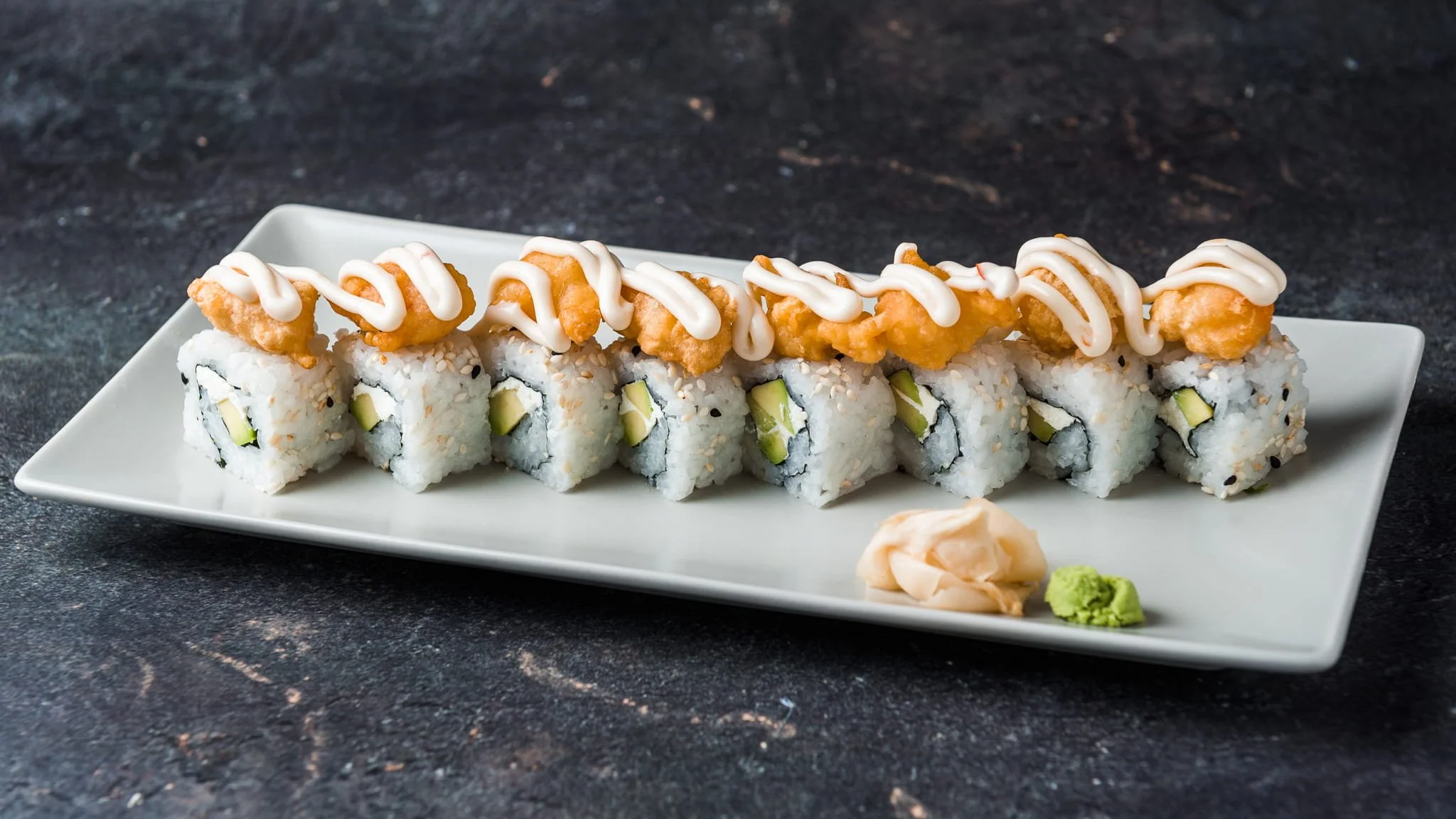 Sushi roll topped with fried chicken pieces, drizzled with white sauce, served with pickled ginger and wasabi on a white rectangular plate.