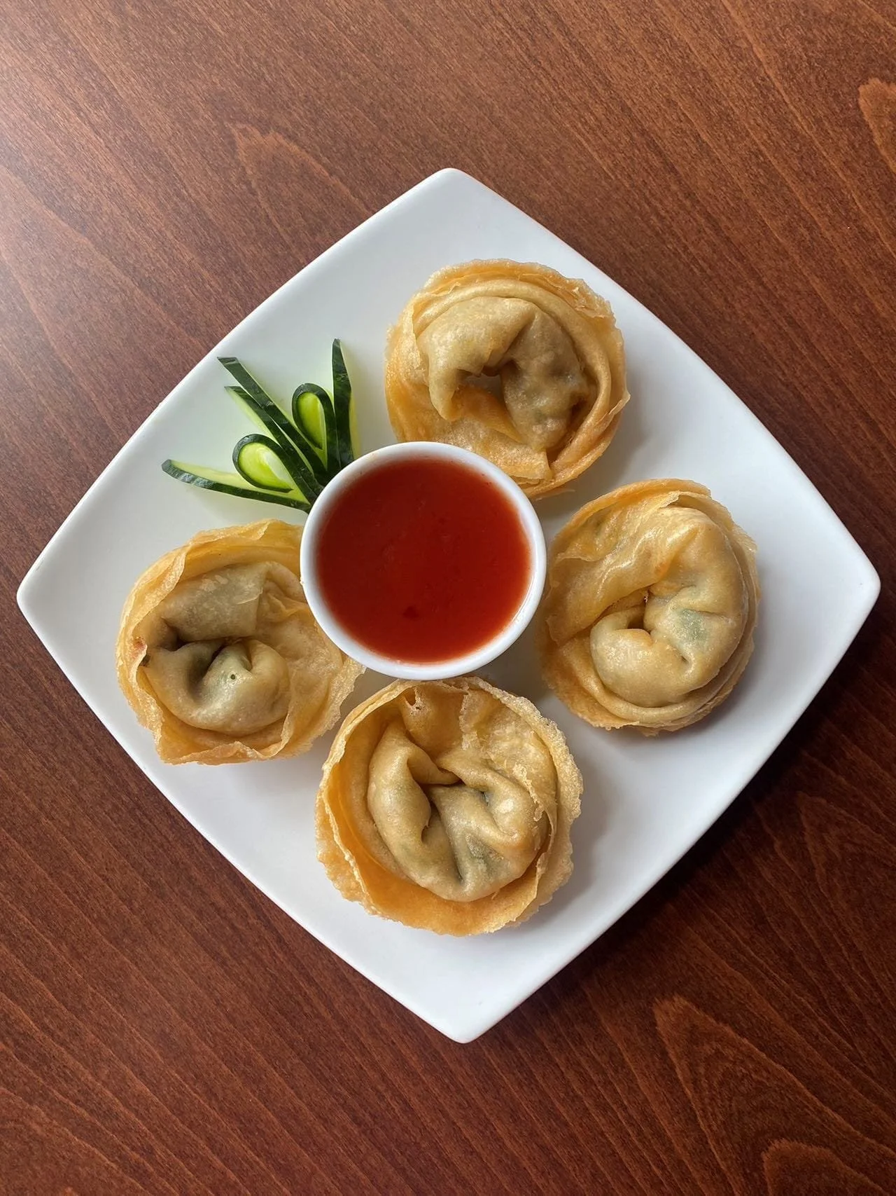 Five fried dumplings with a dipping sauce on a white square plate, garnished with cucumber slices on a wooden table.