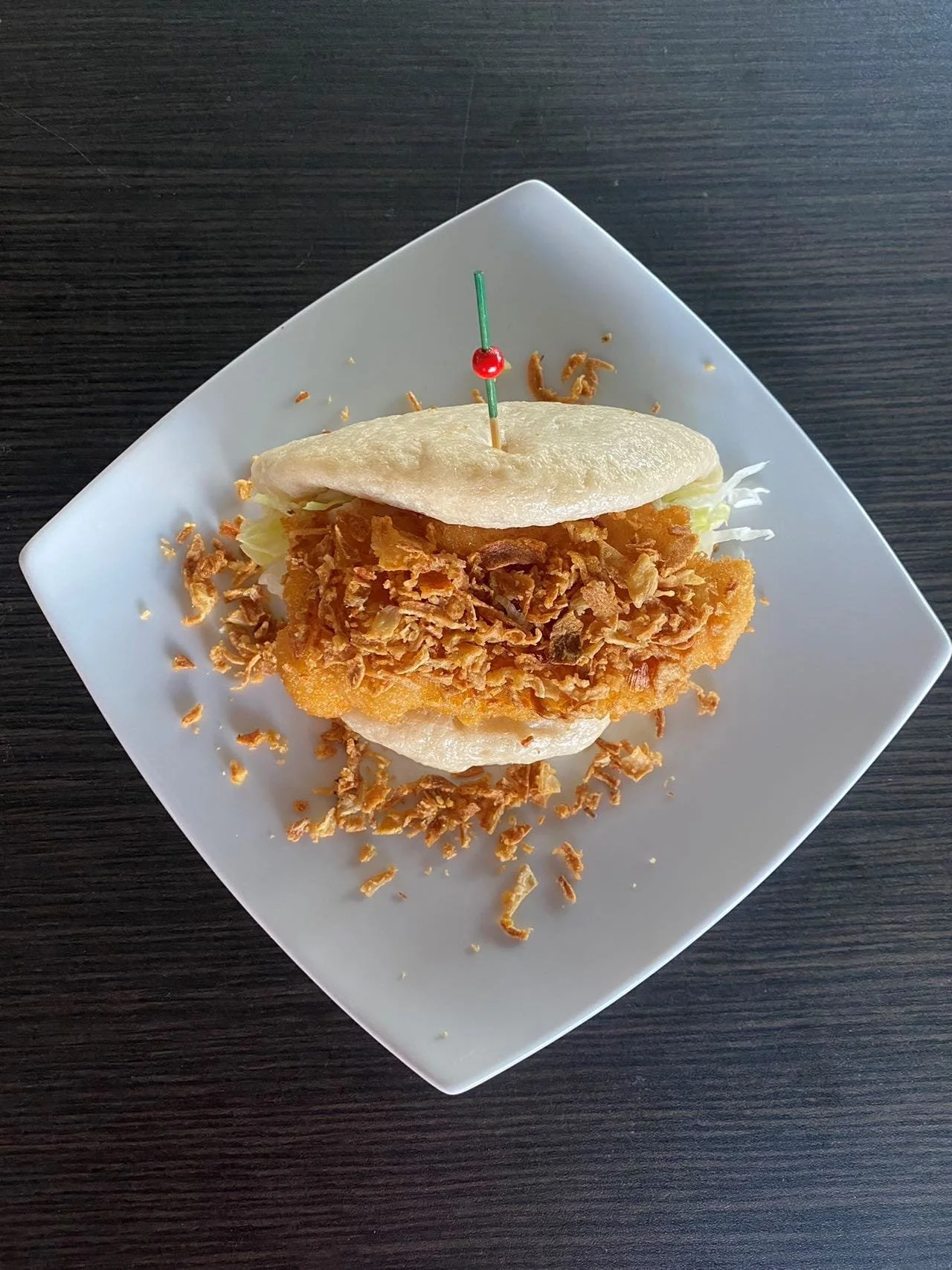 Fried fish sandwich with shredded lettuce on a bun, served on a white square plate with a red toothpick, sitting on a dark wood table.