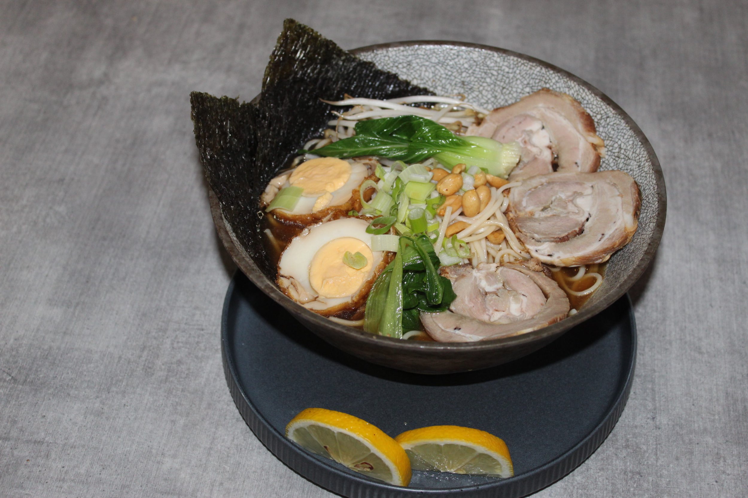 Bowl of ramen with boiled eggs, sliced pork, seaweed, green onions, bean sprouts, and lemon wedges on a black tray.