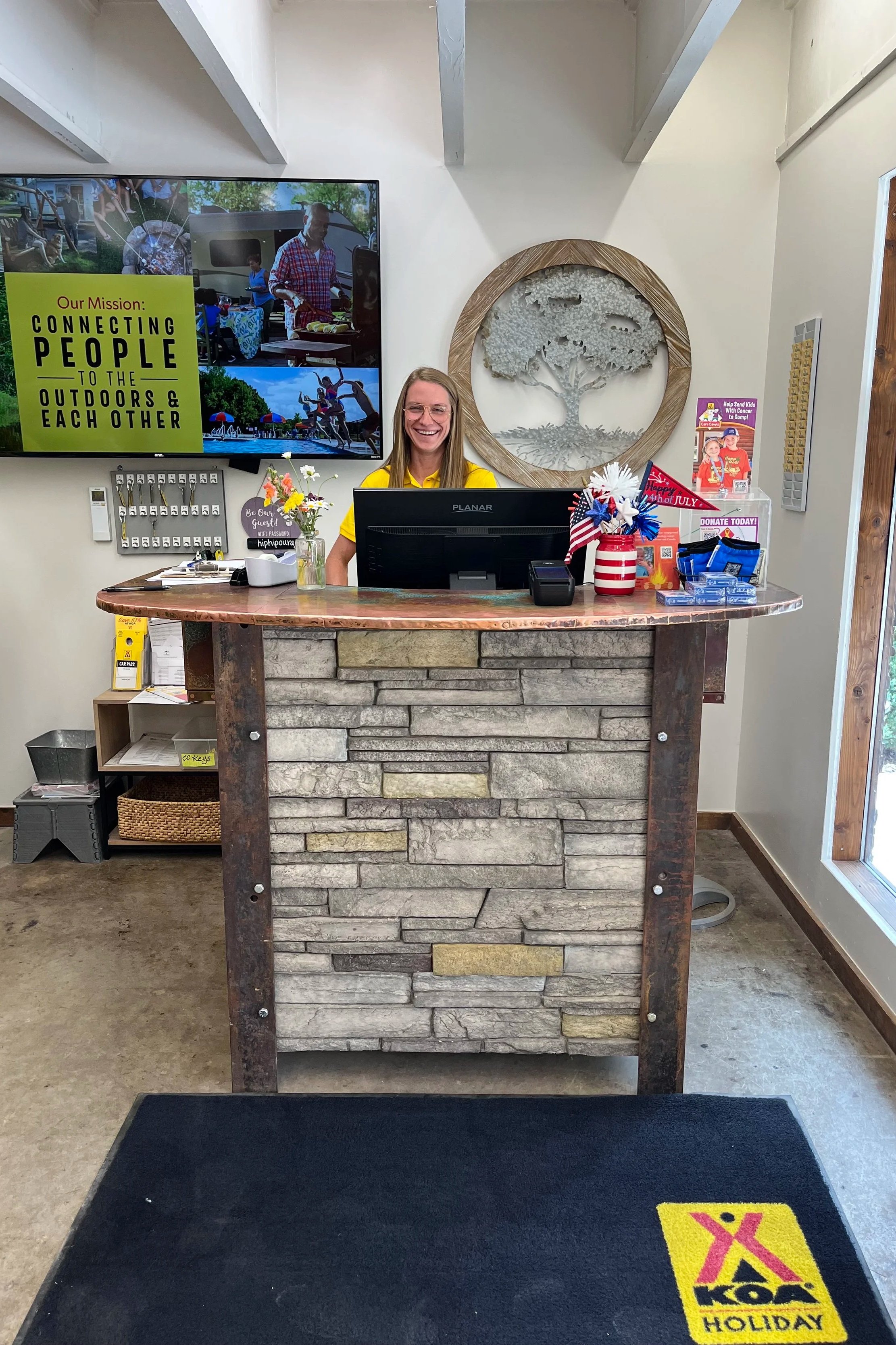A smiling woman standing behind a stone and wood reception desk with a computer. Behind her is a wall-mounted TV screen with a mission statement about connecting people with the outdoors. There are decorations and informational signs on the desk, and sunlight coming through a window to the right.