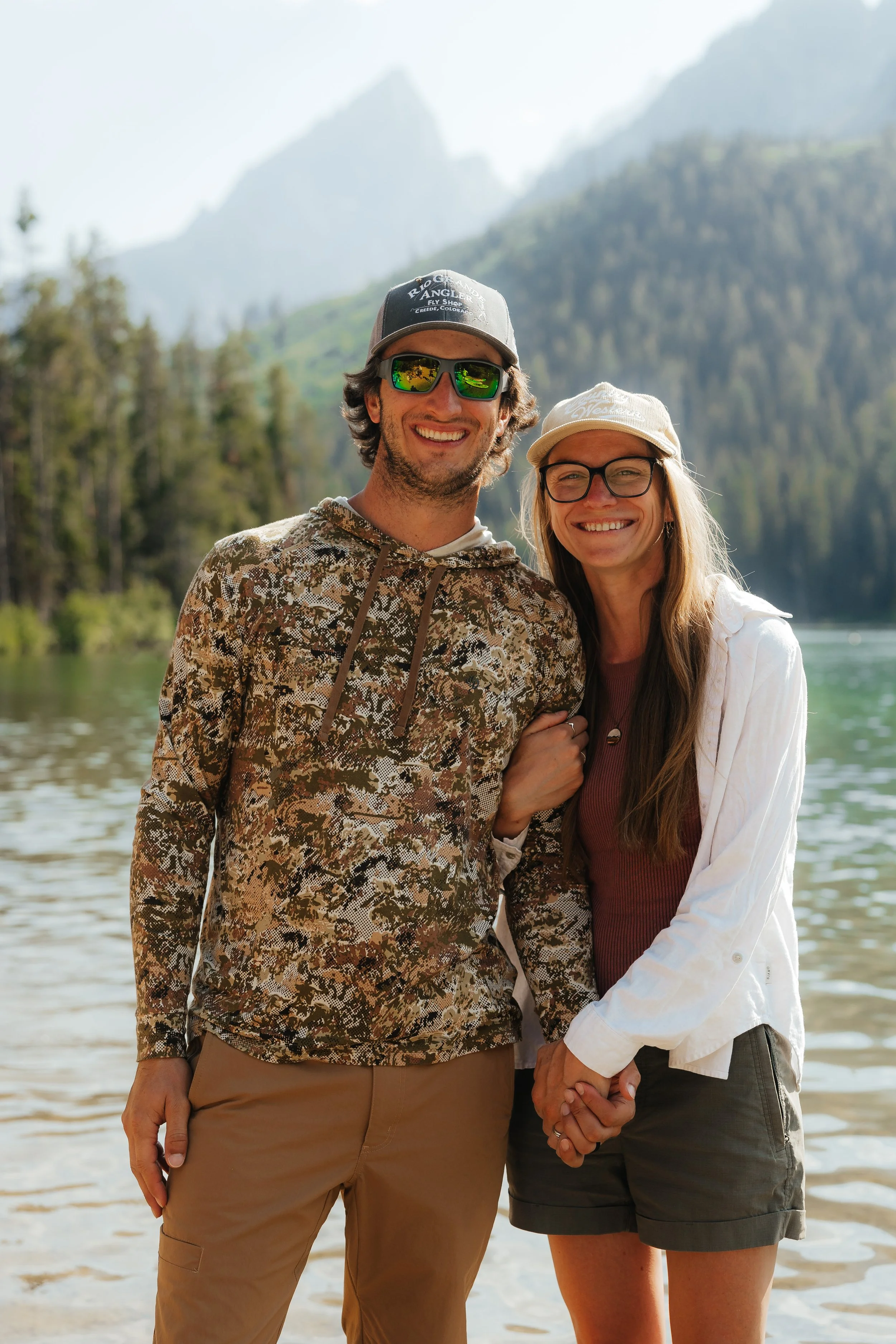 A happy couple holding hands by a lake with mountains and trees in the background.