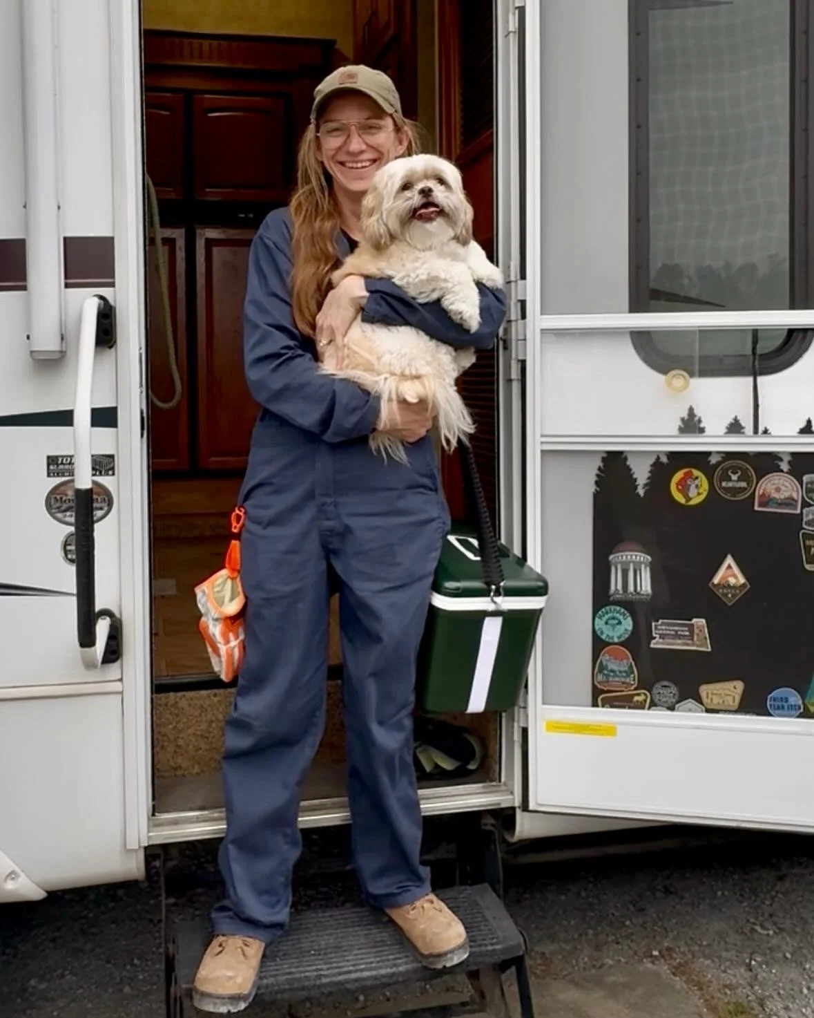 A woman in construction coveralls holding a small fluffy dog at the entrance of a camper, with stickers on the door.