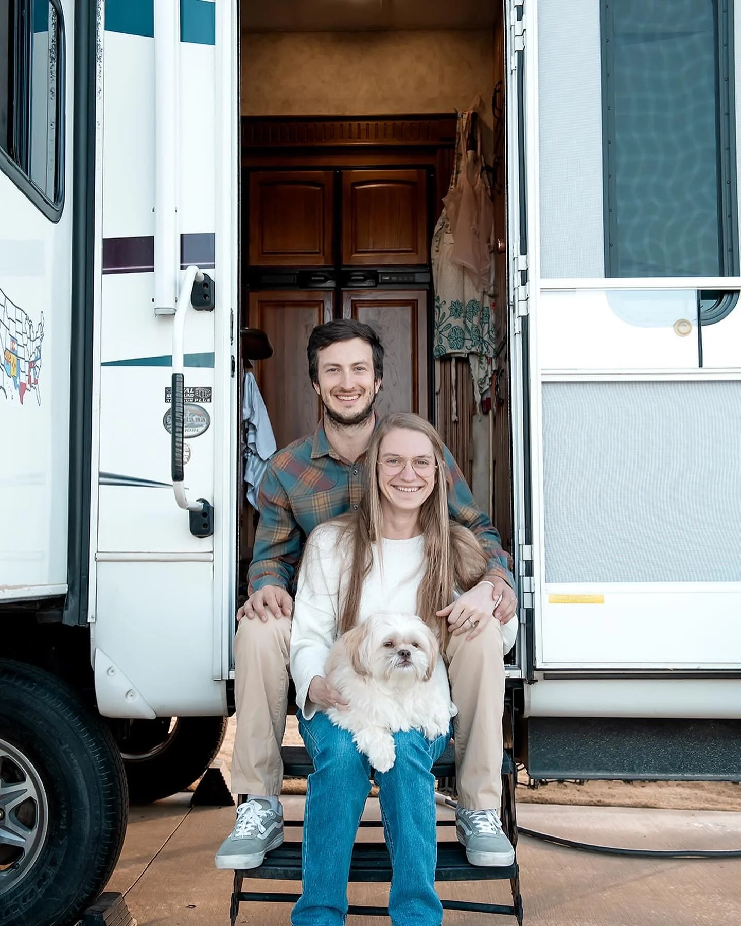 A smiling couple sitting on the steps of a camper with their small white dog, outdoors on a clear day.