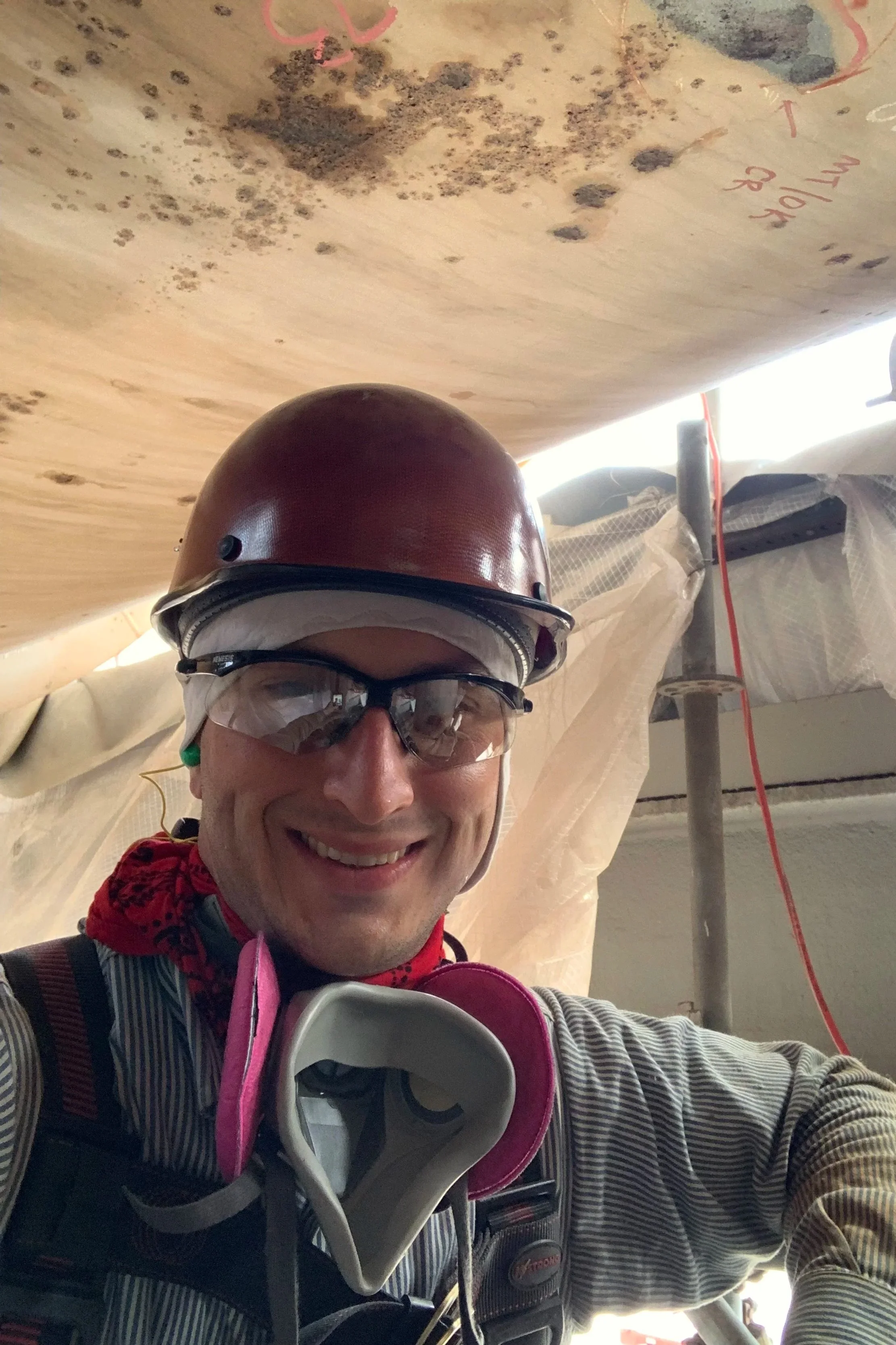 A man wearing a construction helmet, safety goggles, and a mask with pink filters taking a selfie indoors with wooden beams and construction materials in the background.