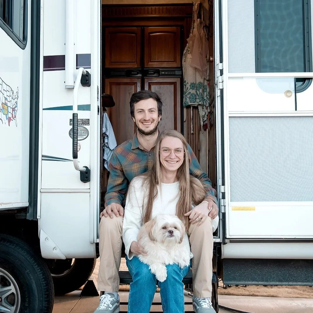 A smiling couple sitting in front of a camper, with the woman holding a small dog.