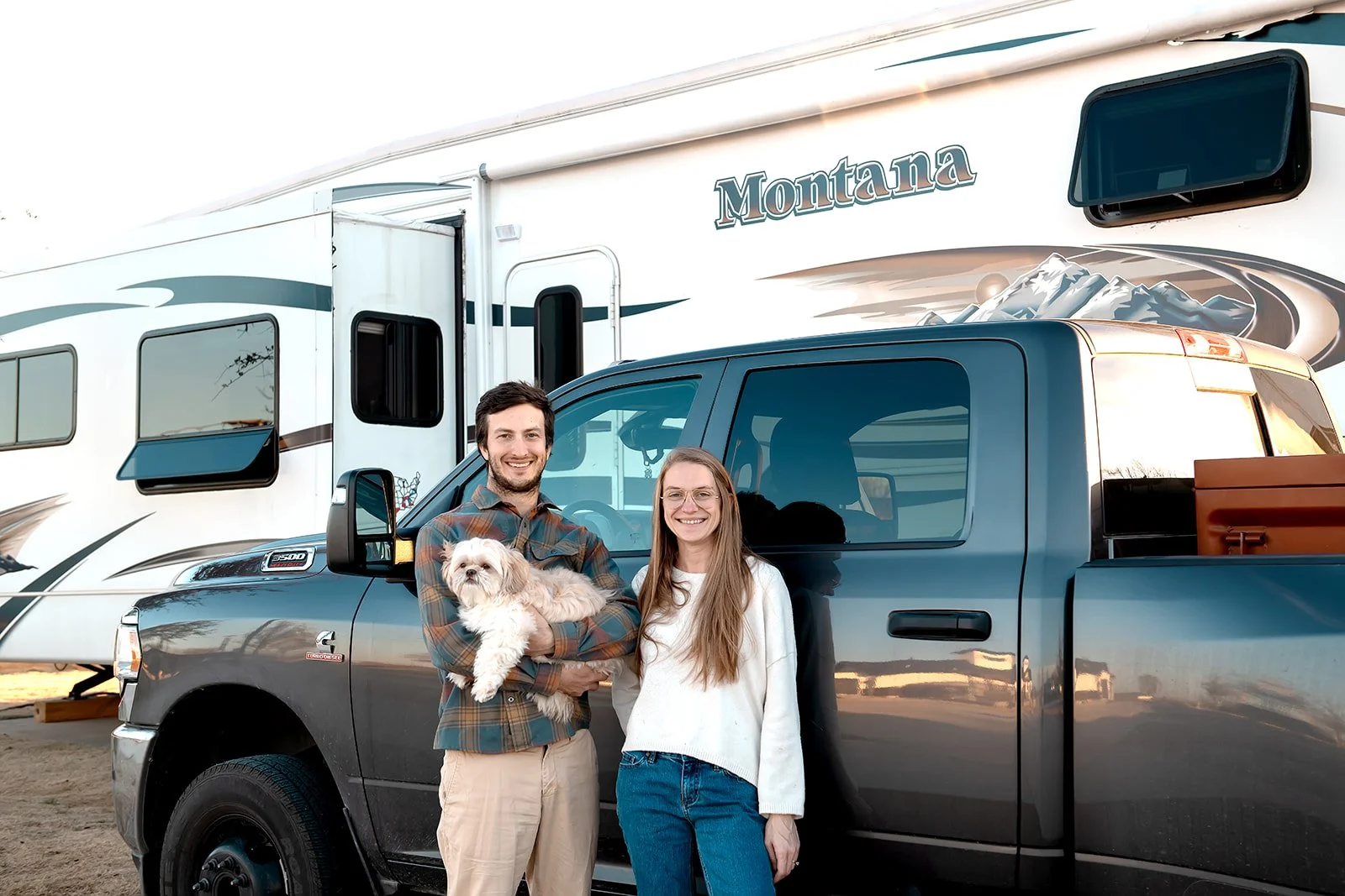 A smiling man and woman standing next to a dark gray pickup truck, with the man holding a small white dog. Behind them is a white and teal Montana fifth-wheel camper. The scene appears to be outdoors during the daytime.