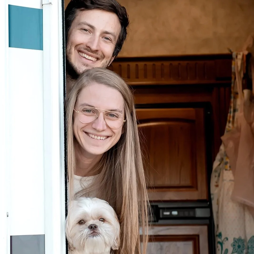 A smiling couple with a small white dog peeking out from a doorway inside a RV.