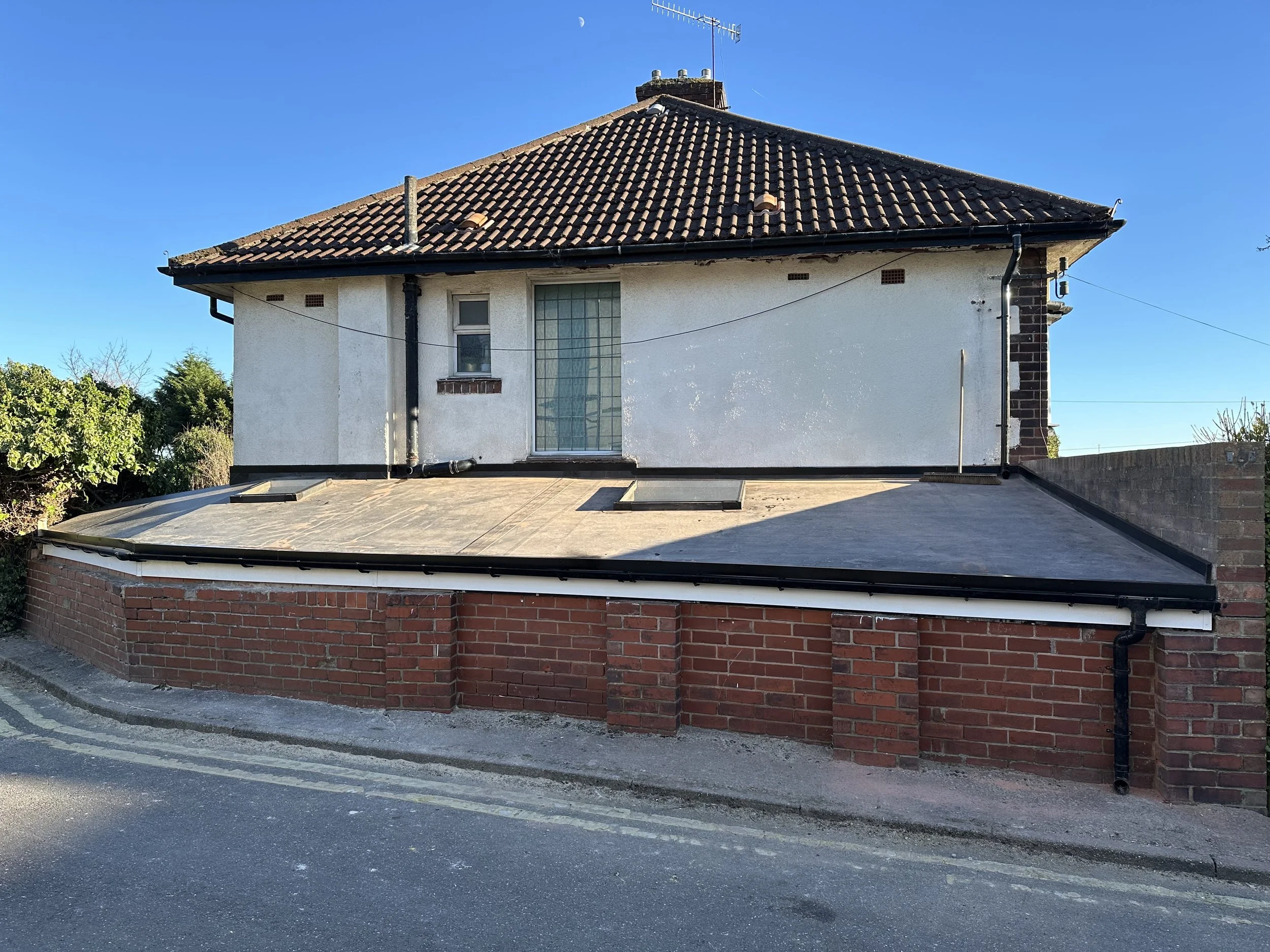 A two-story house with a tiled roof, white walls, a large glass block window, and a flat roof extension with a brick base, situated at the corner of a street under a clear blue sky.