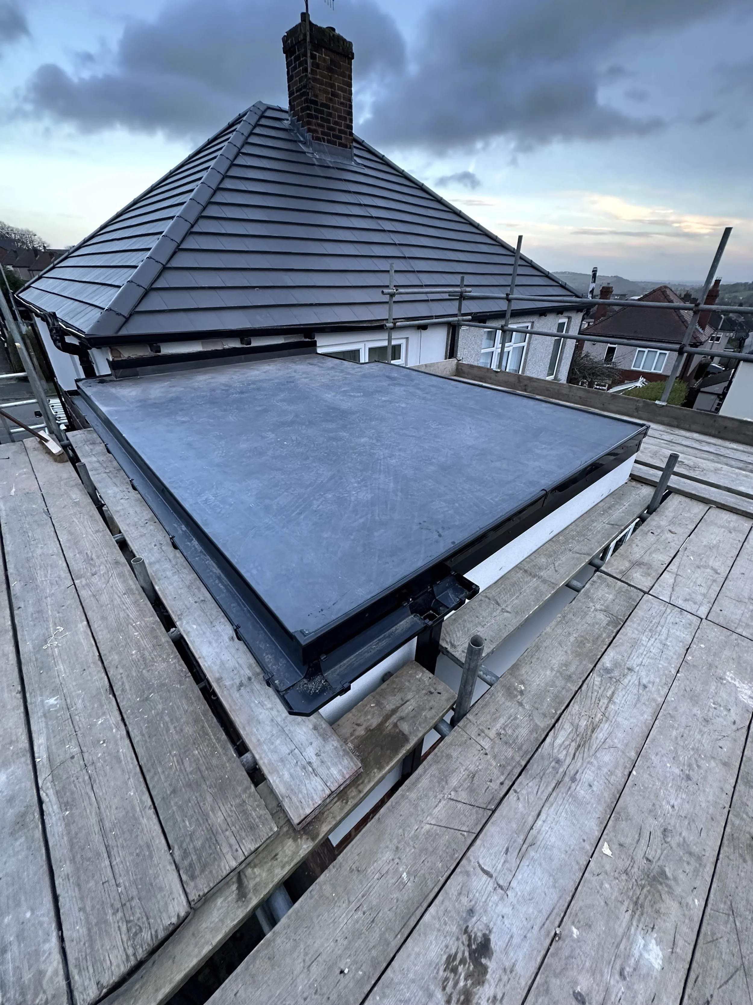Roof of a house under construction with scaffolding, new flat roof section, and surrounding residential homes, cloudy sky in the background.