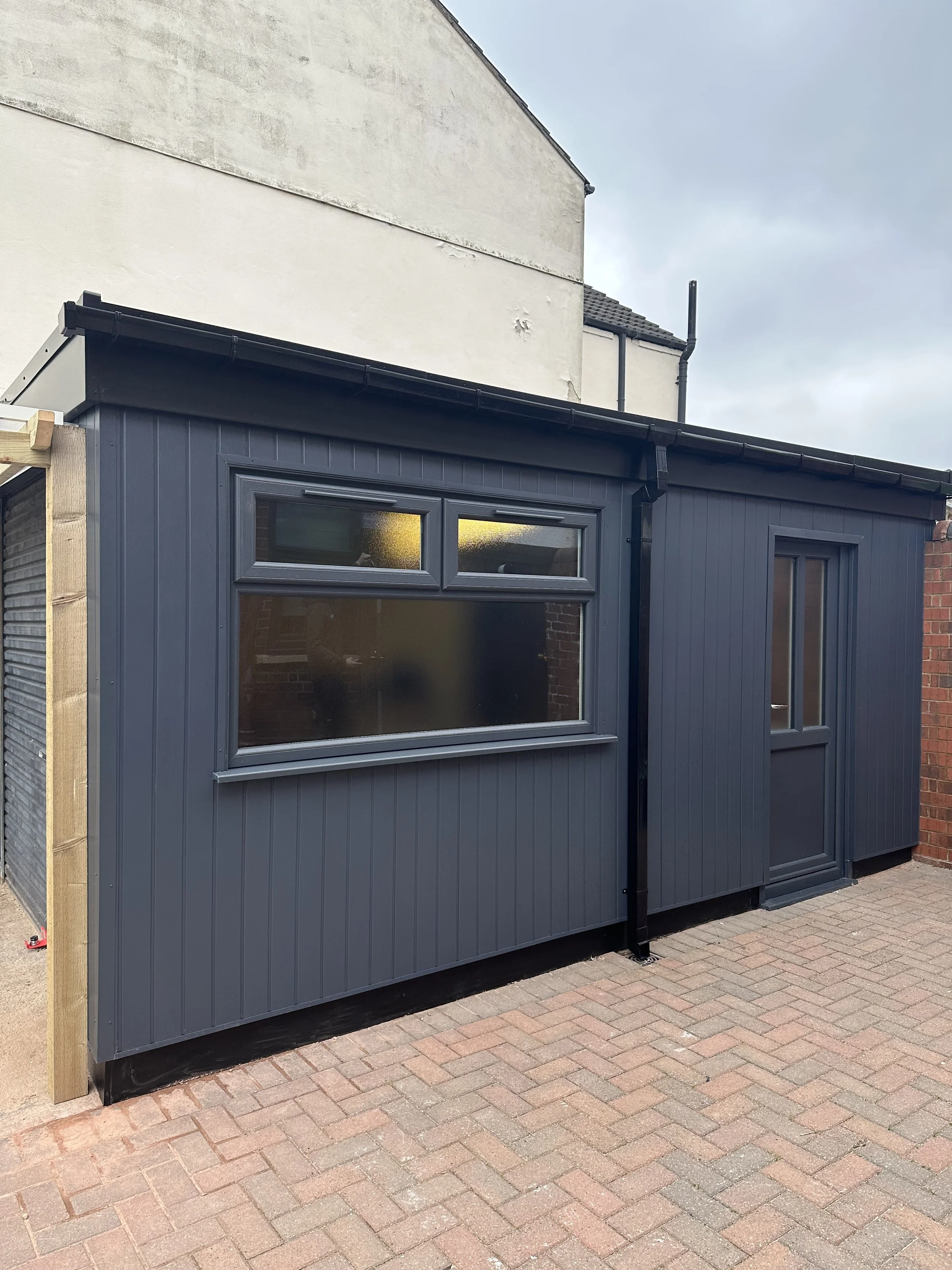 Modern dark gray garden shed with a window, a door, and vertical siding, situated on a brick patio.