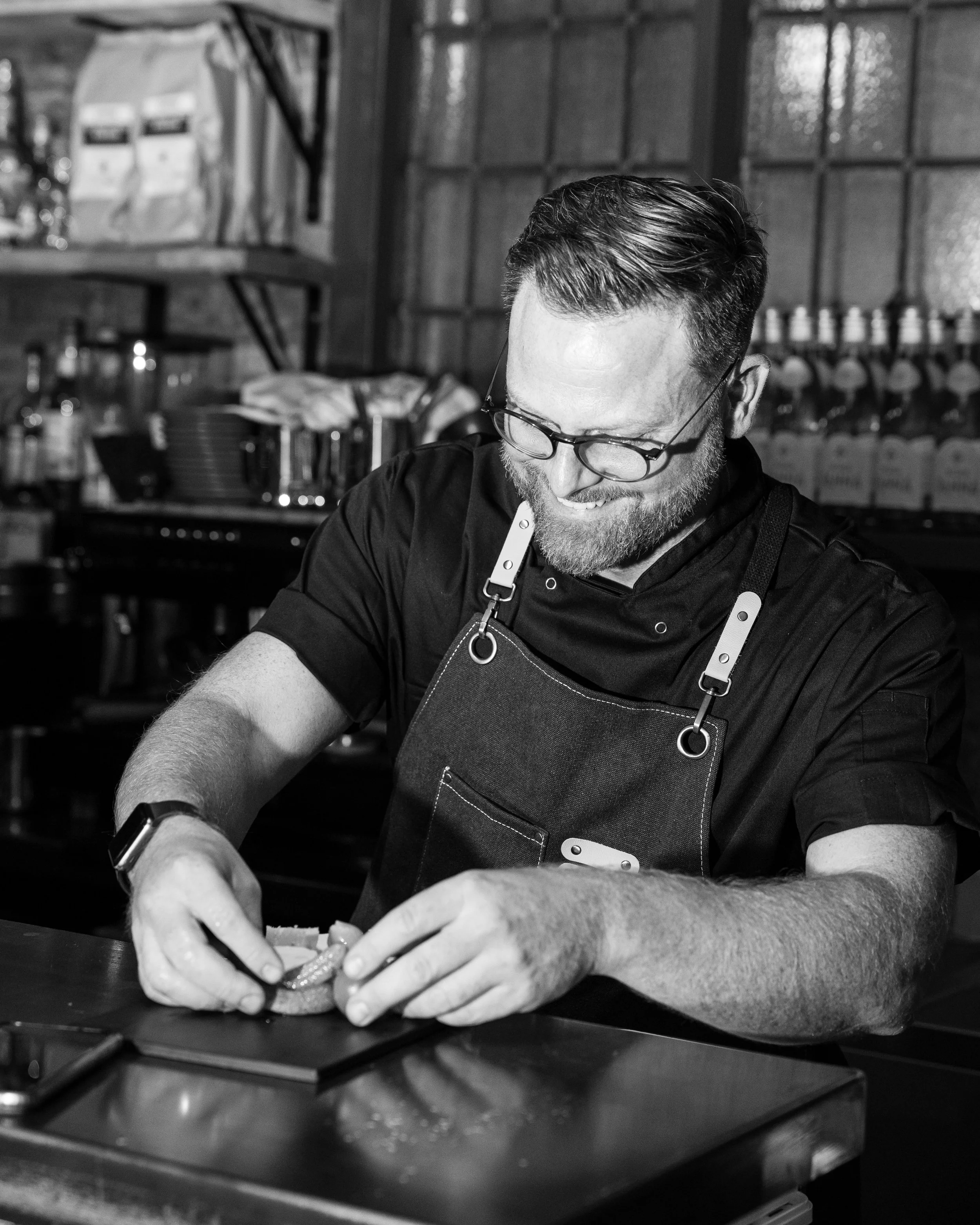 A man wearing glasses, a black apron, and a black shirt, smiling as he prepares food in a kitchen.