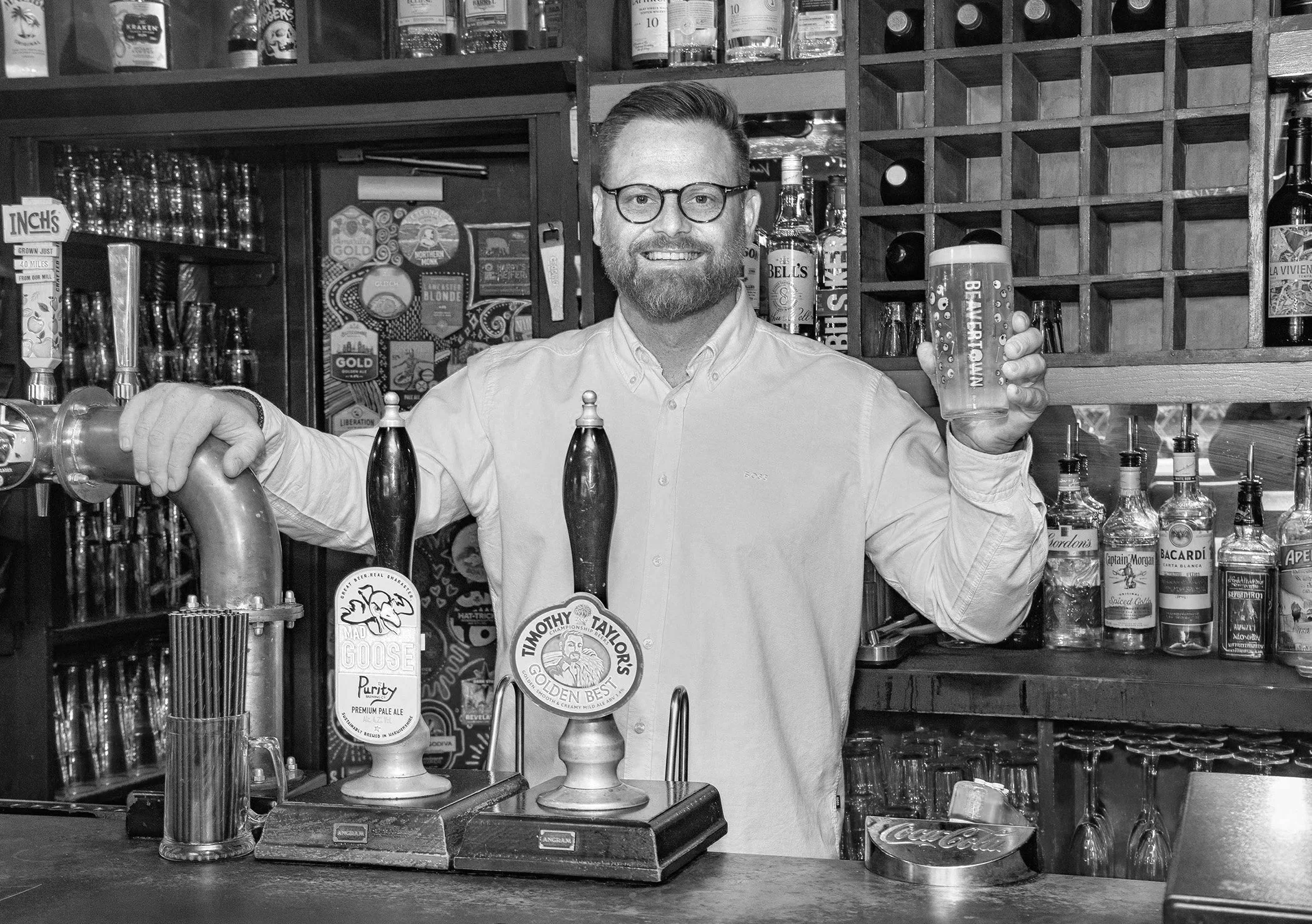 Matt Crowther behind the bar at The Black Pug Warwick, smiling, holding a pint of beer in his right hand. The bar has various bottles of alcohol and beer taps in front of him.