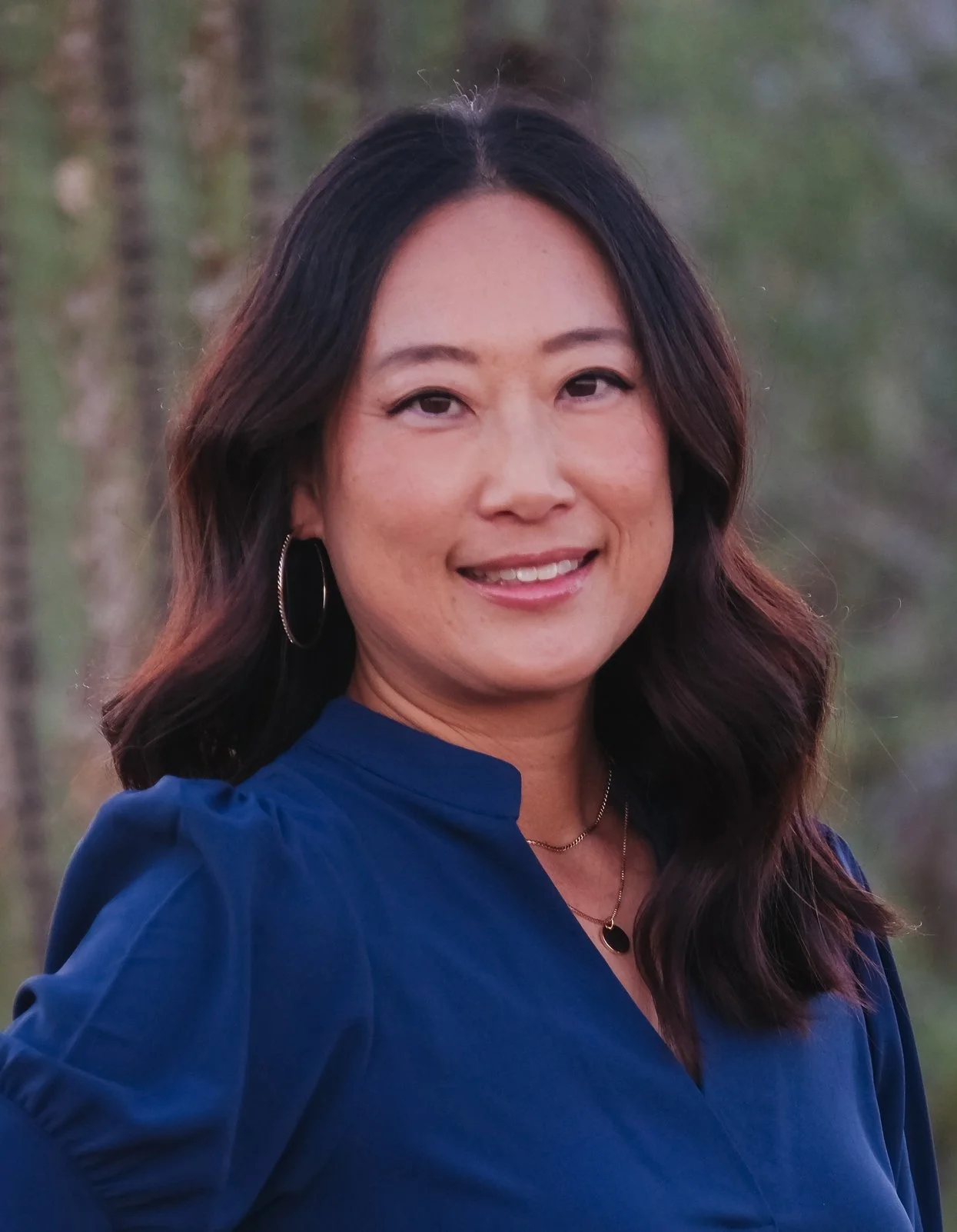 A woman with dark brown hair, wearing a blue top, earrings, and a necklace, smiling outdoors with a background of trees.