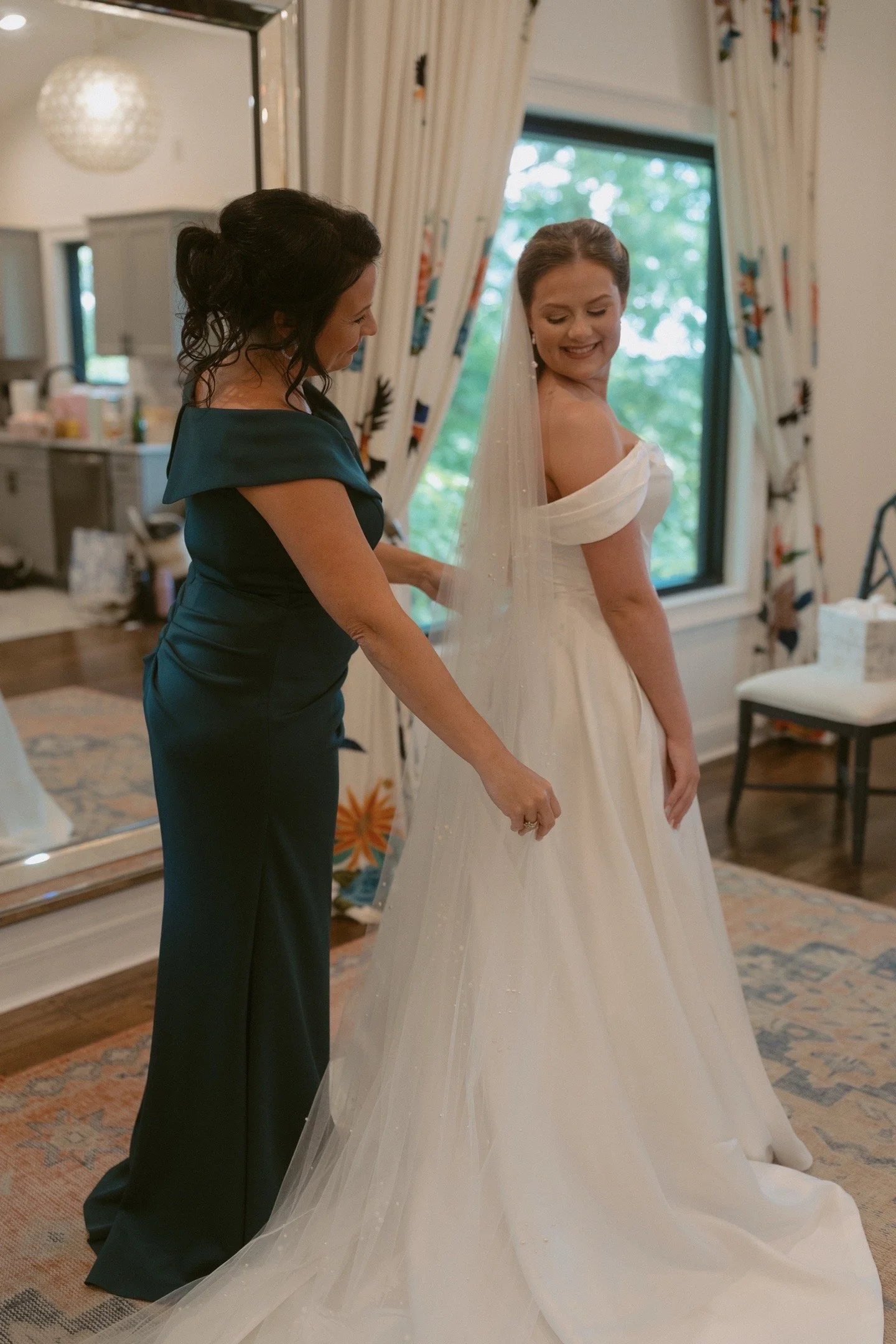 A bride in a white wedding gown smiling as she looks at herself in a mirror, with a woman helping her adjust her dress in a room with a large window and floral curtains.