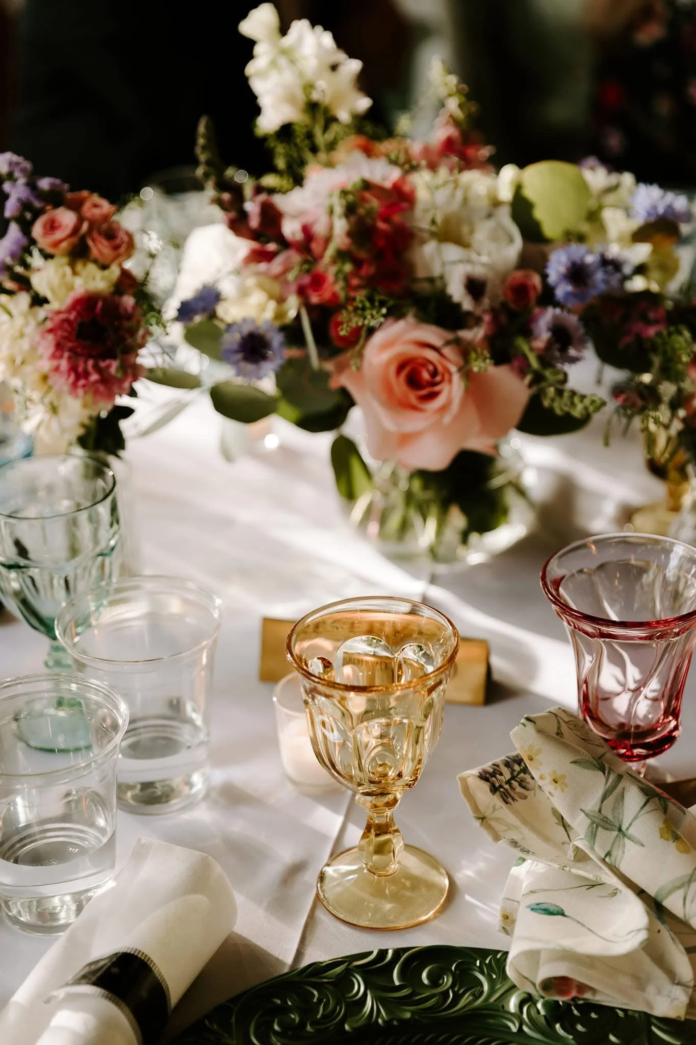 A close-up of a set dining table with a floral centerpiece, wine glasses, water glasses, and napkins. The centerpiece features pink roses, purple, white, and red flowers with greenery. The setting appears elegant and decorative, suitable for a specia