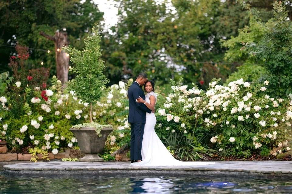 A bride and groom standing close together by a pool, surrounded by lush green trees and blooming white flowers in a garden setting.