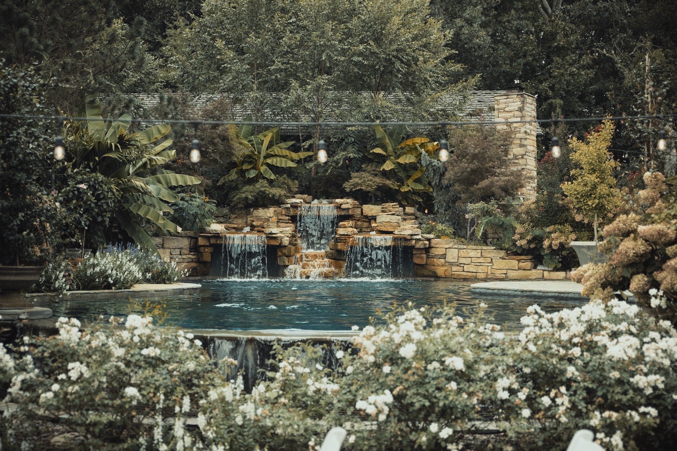 A landscaped backyard with a pond, a small waterfall made of stone, surrounded by lush greenery and flowering plants, with string lights hanging overhead.