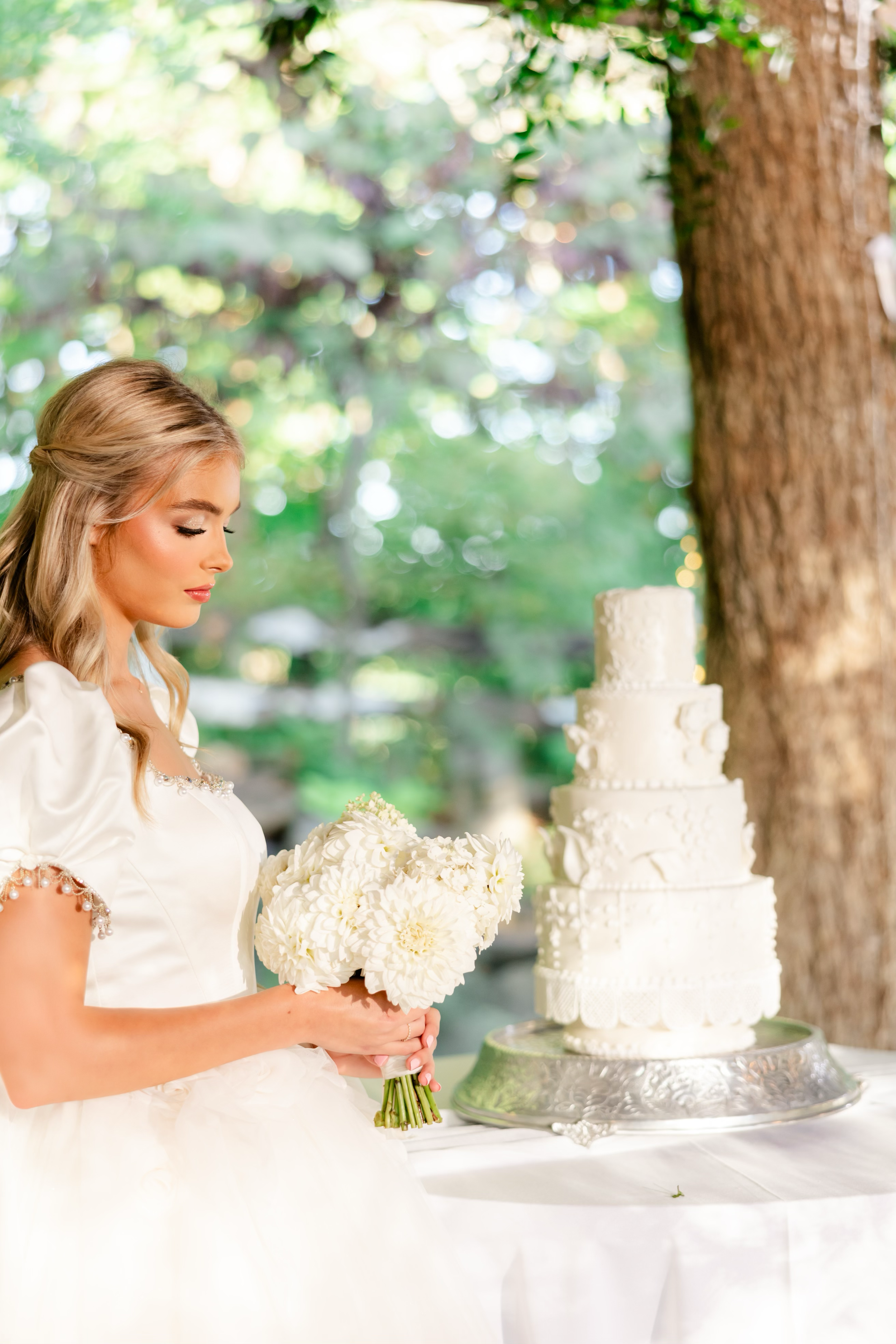 A bride in a white wedding dress standing beside a tall white wedding cake at an outdoor reception, holding a bouquet of white flowers, with a tree and lush greenery in the background.