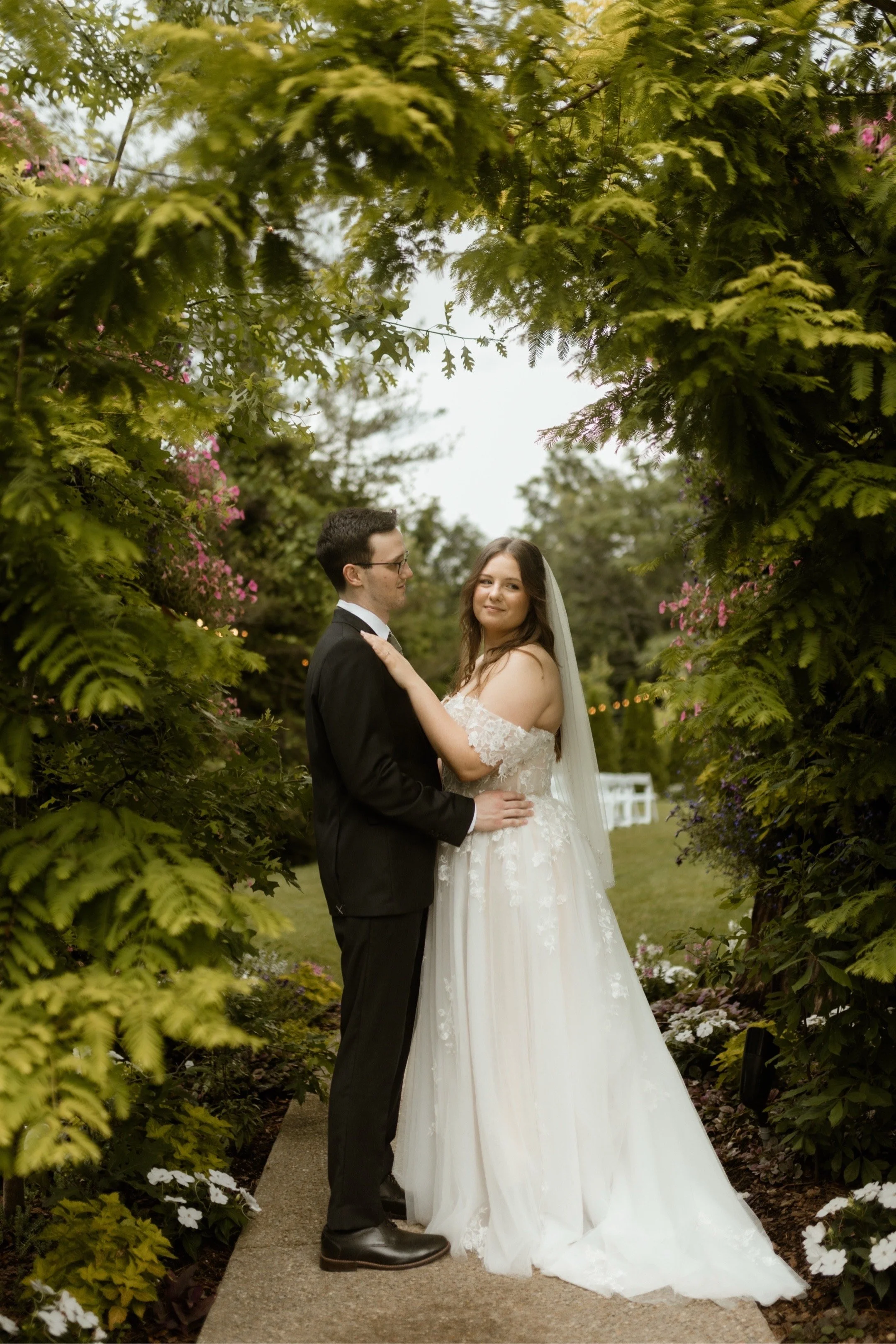 A bride and groom pose together outdoors on a wedding day, surrounded by lush greenery and blooming flowers, with a garden pathway visible.
