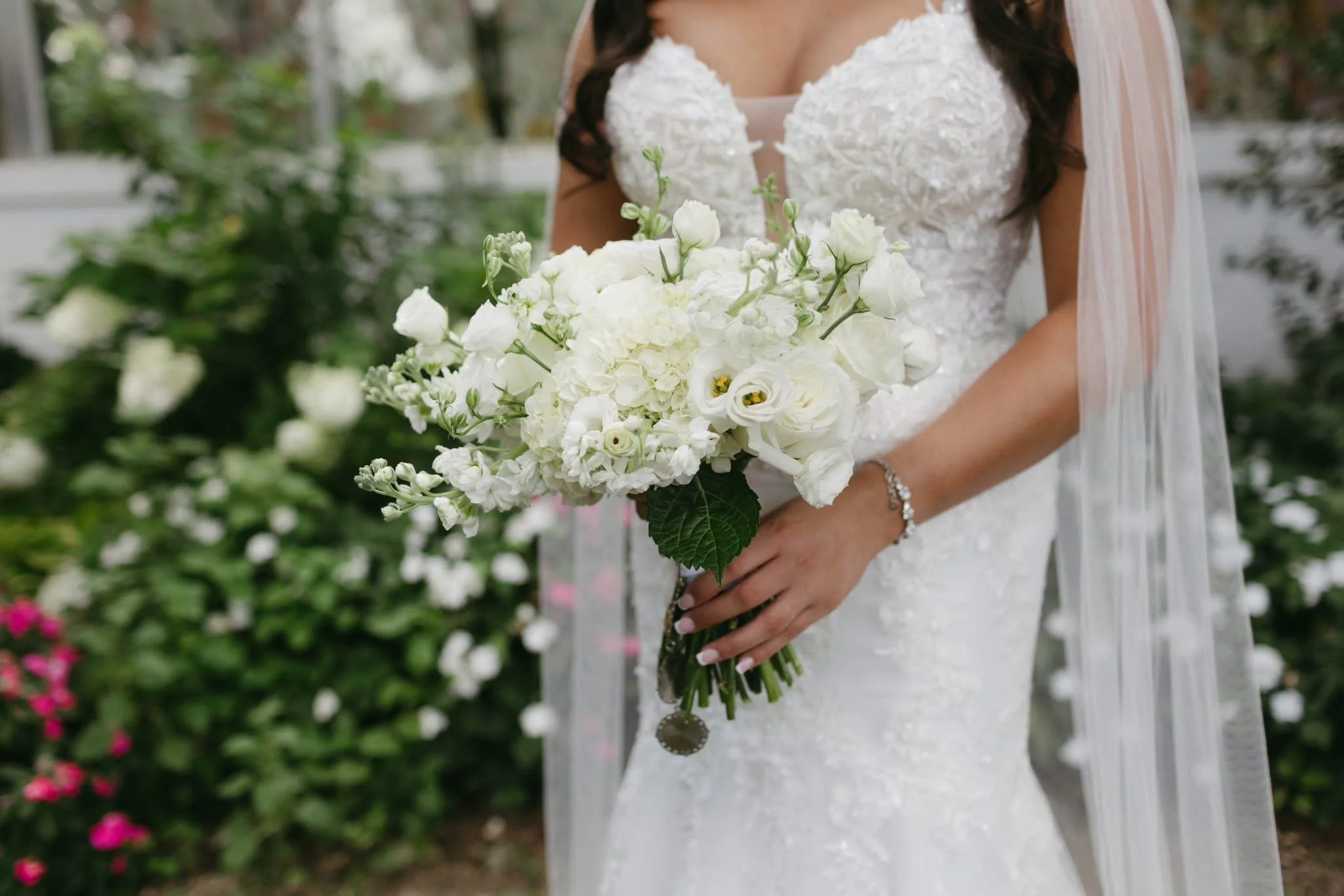 Bride holding a bouquet of white flowers in a garden setting, wearing a white lace wedding dress and veil.