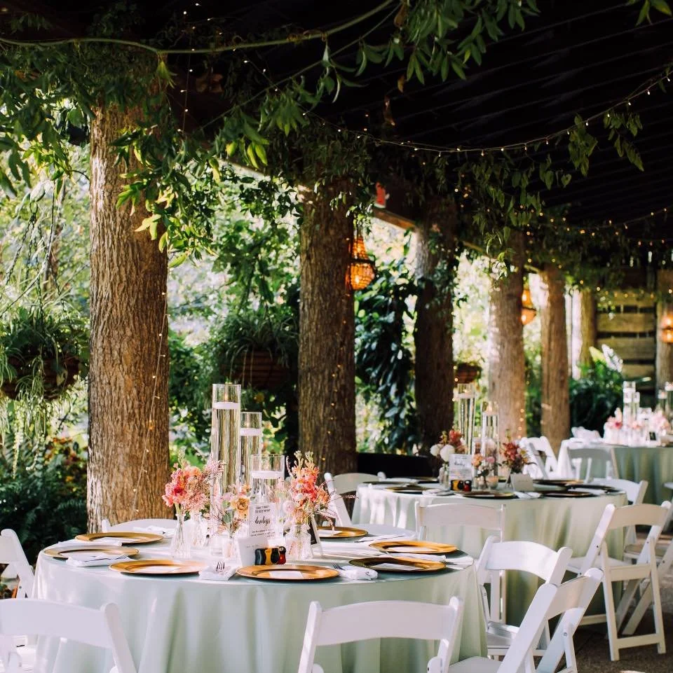 Outdoor event setup with round tables, white chairs, floral centerpieces, candles, and string lights hanging from trees.