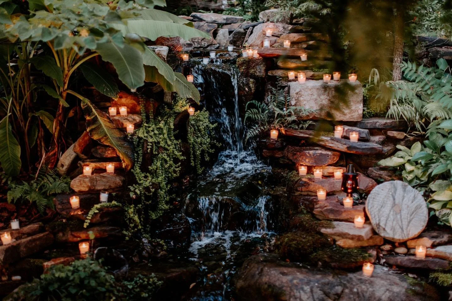A small waterfall surrounded by lush green plants and rocks, with numerous lit candles placed along the rocks and around the waterfall creating a serene and cozy ambiance.