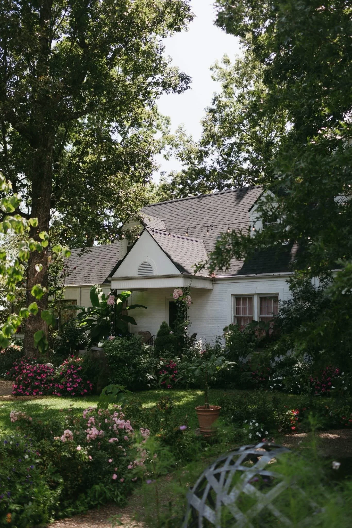 A white house with a gray roof, surrounded by a lush garden with pink and white flowers and green trees, under a partly cloudy sky.