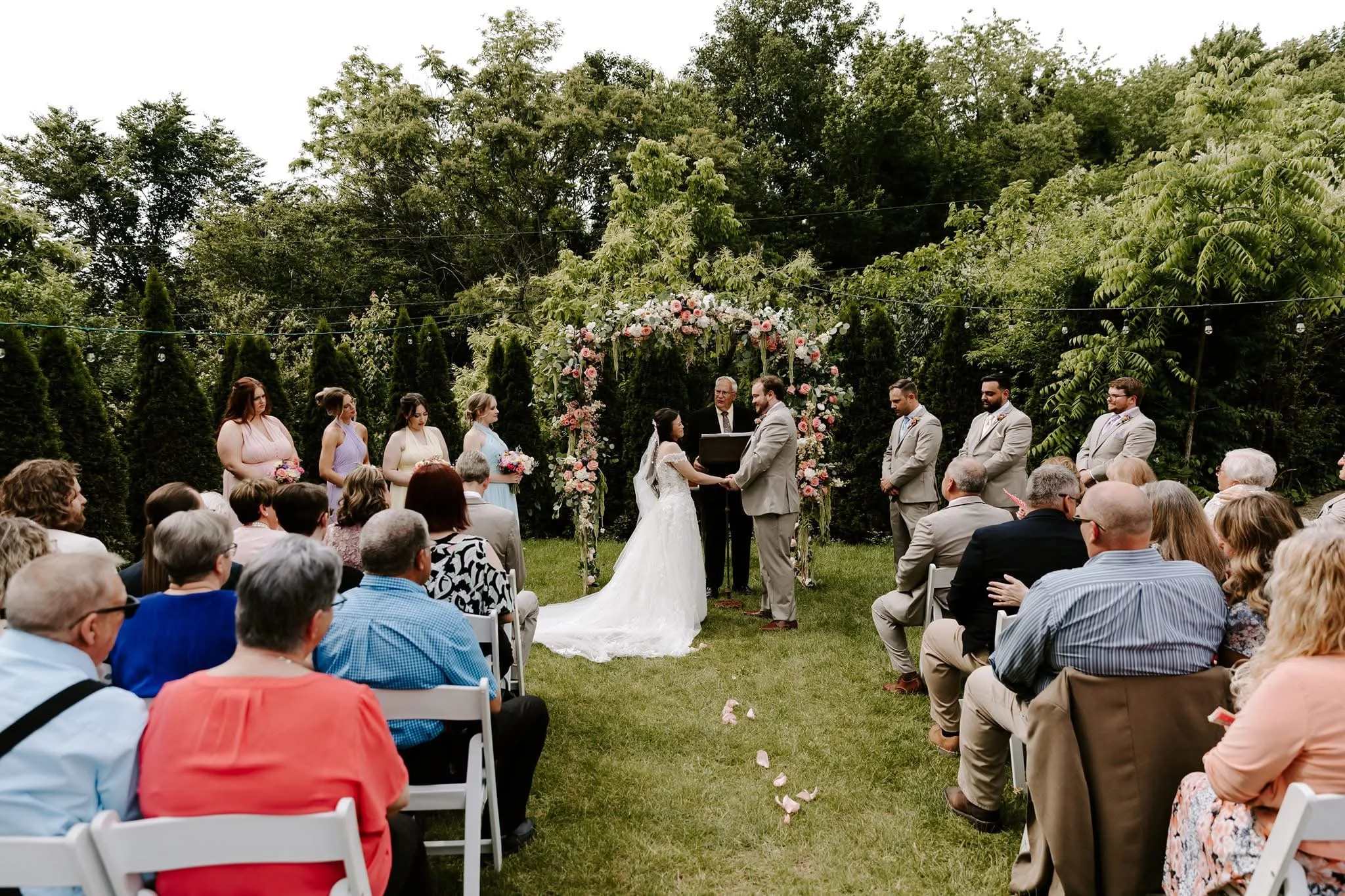 A wedding ceremony taking place outdoors with a bride and groom exchanging vows under a floral arch, surrounded by guests seated on white chairs, officiant, bridesmaids, and groomsmen in suits, greenery backdrop, and overcast sky.