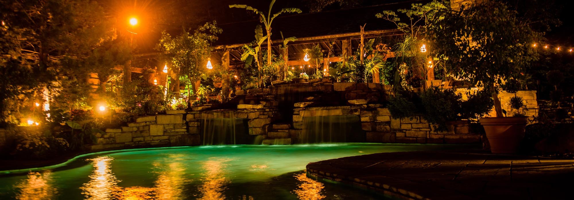Nighttime view of a backyard with a swimming pool, waterfall feature, stone wall, lush plants, and string lights illuminating the area.