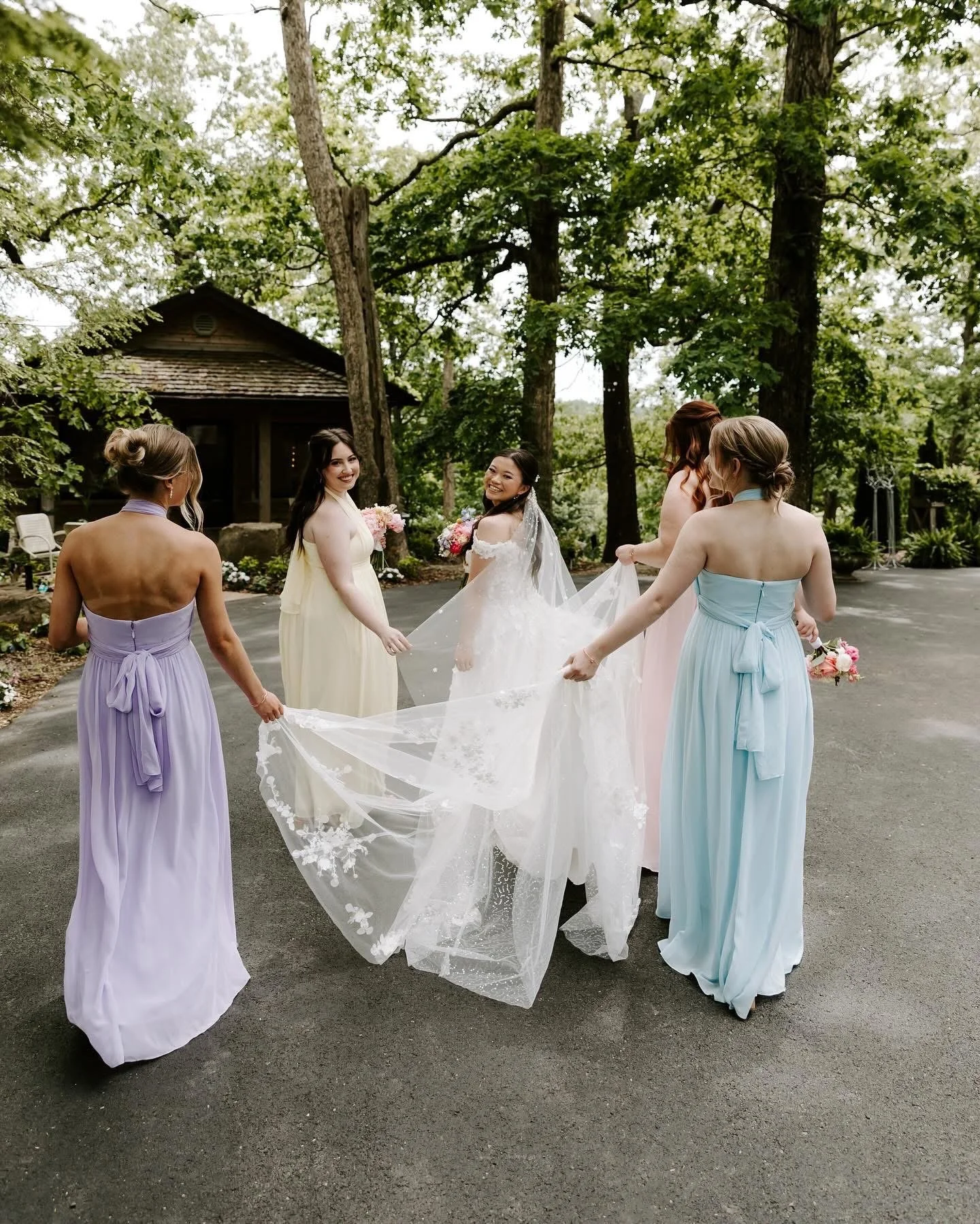 A bride in a white wedding dress holding a bouquet, surrounded by bridesmaids in pastel dresses, holding her train, outdoors among tall trees.