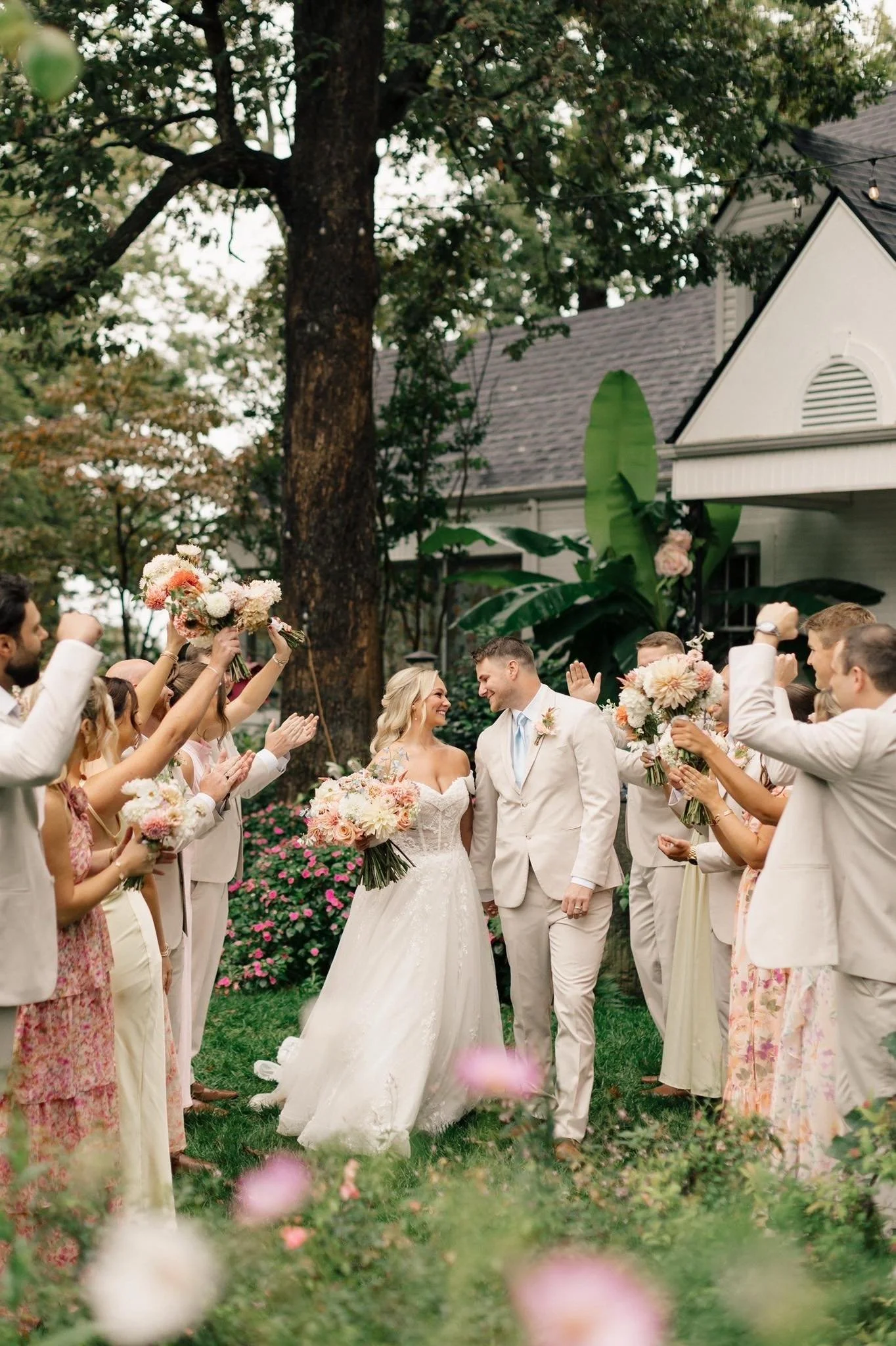 A bride and groom walking together under an arch of flowers, surrounded by friends and family celebrating during a wedding ceremony outdoors.