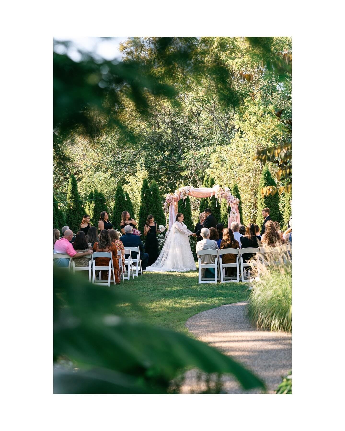 A wedding ceremony outdoors in a lush green garden with trees and shrubs. The bride and groom are holding hands at the altar decorated with pink and white flowers, surrounded by guests seated on white chairs.