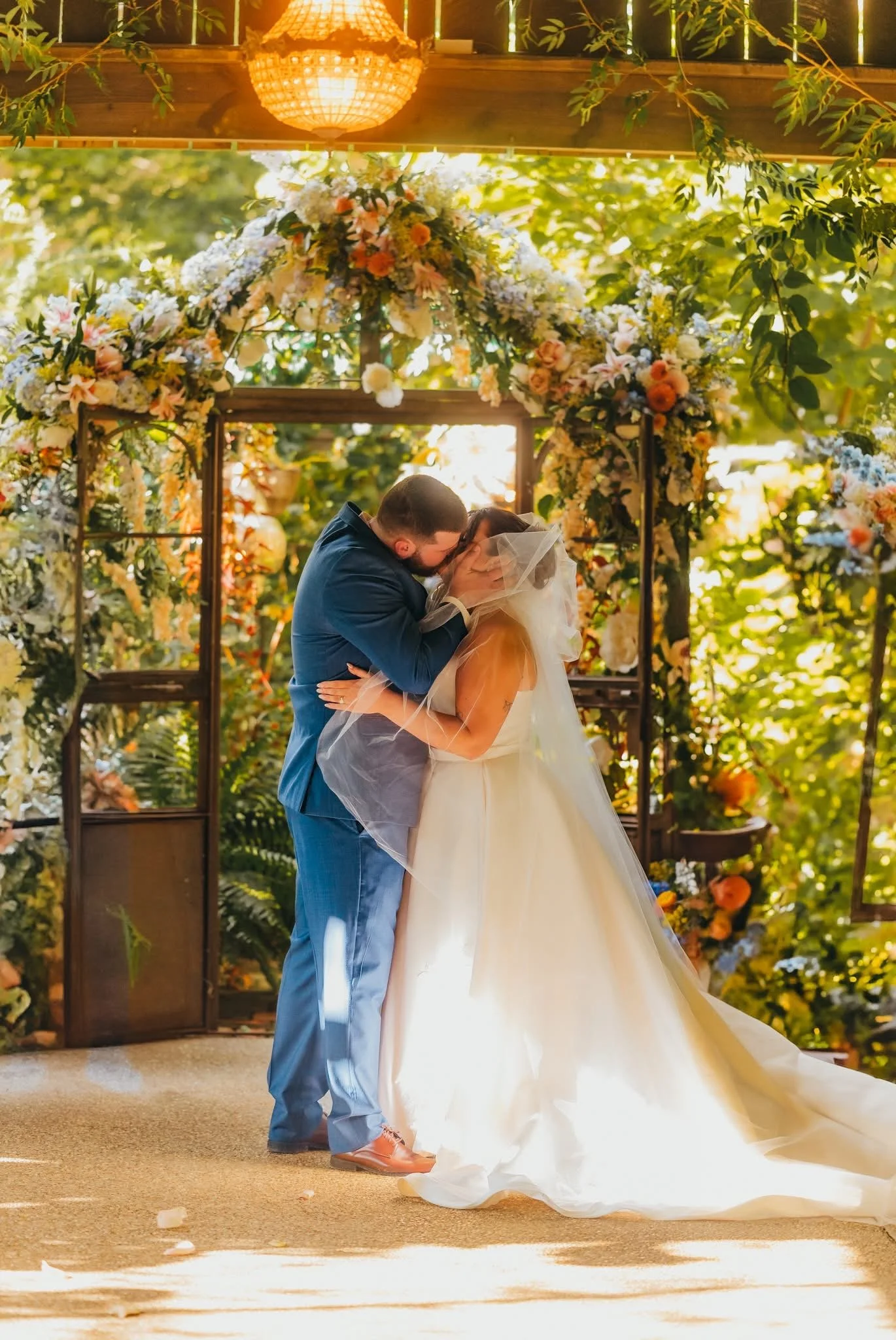 A bride and groom are kissing during their wedding ceremony in an outdoor setting decorated with flowers and greenery.