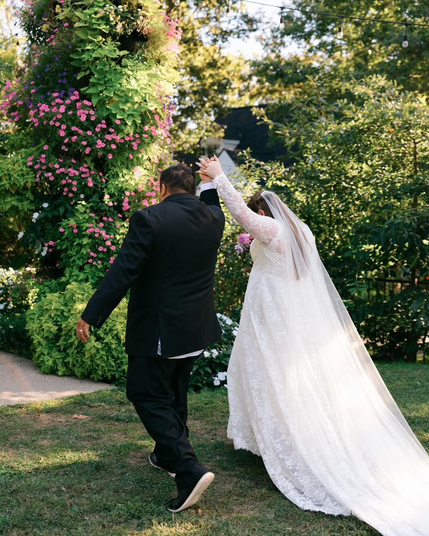 A bride and groom dancing outdoors in a garden, surrounded by lush green foliage and pink flowers, with sunlight filtering through trees.