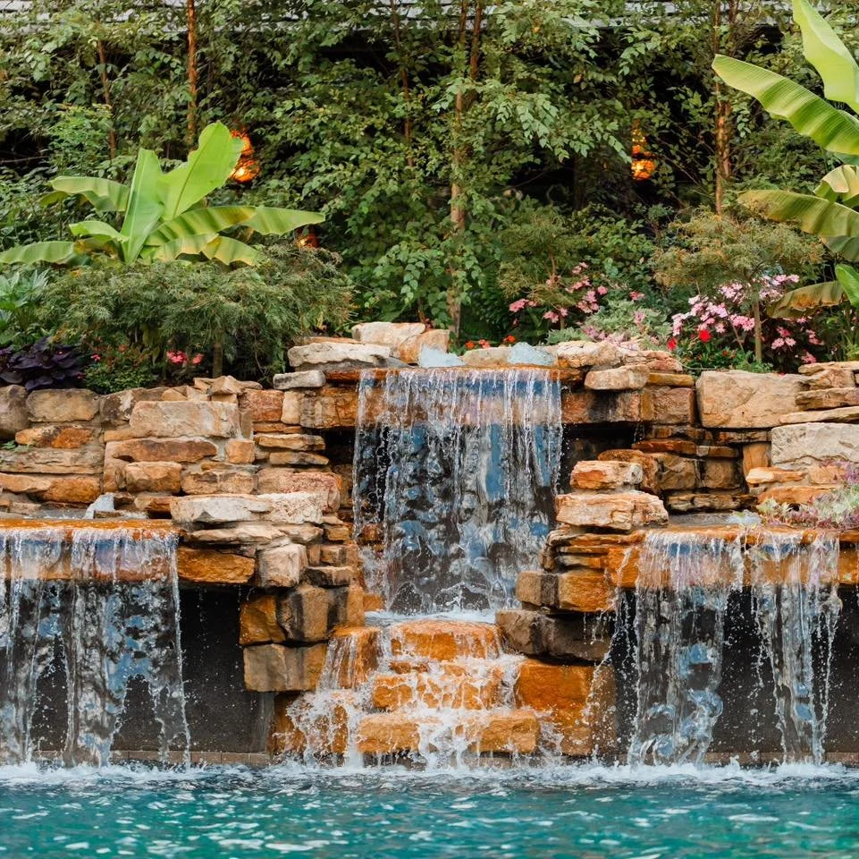 Waterfall cascading over rocks into a pool, surrounded by lush greenery and flowering plants.