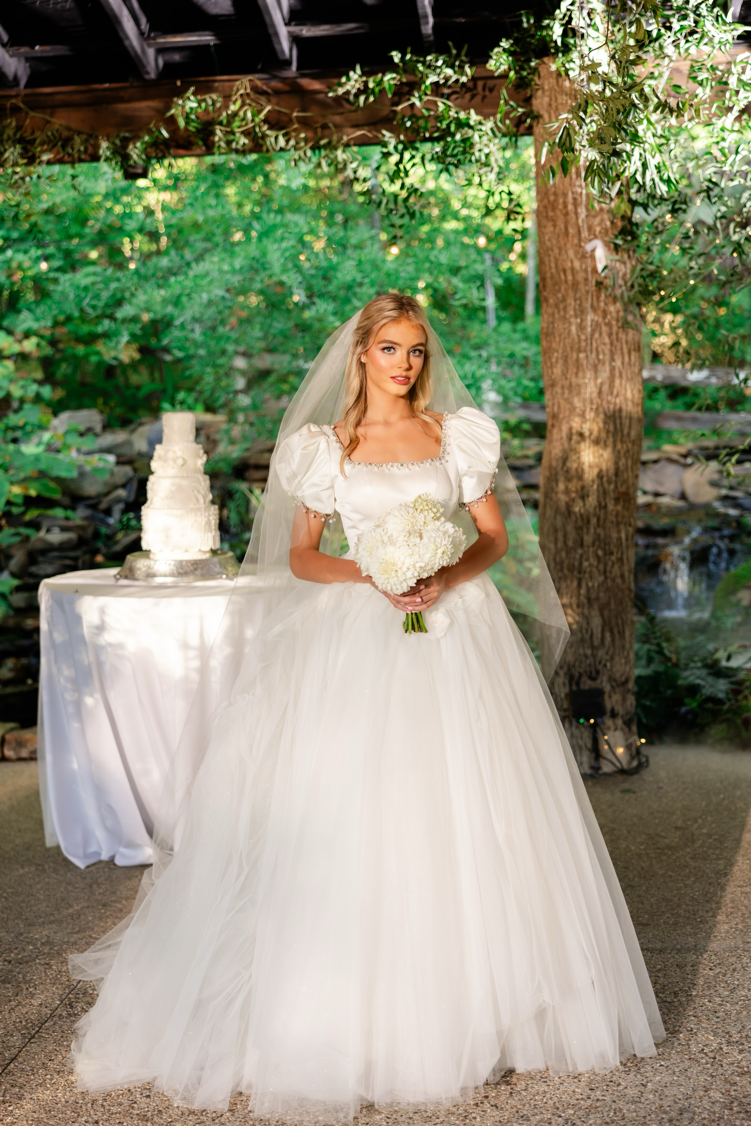 A bride in a white wedding gown holding a bouquet of white flowers standing outdoors in front of a table with a white wedding cake, under a wooden structure with greenery and string lights.