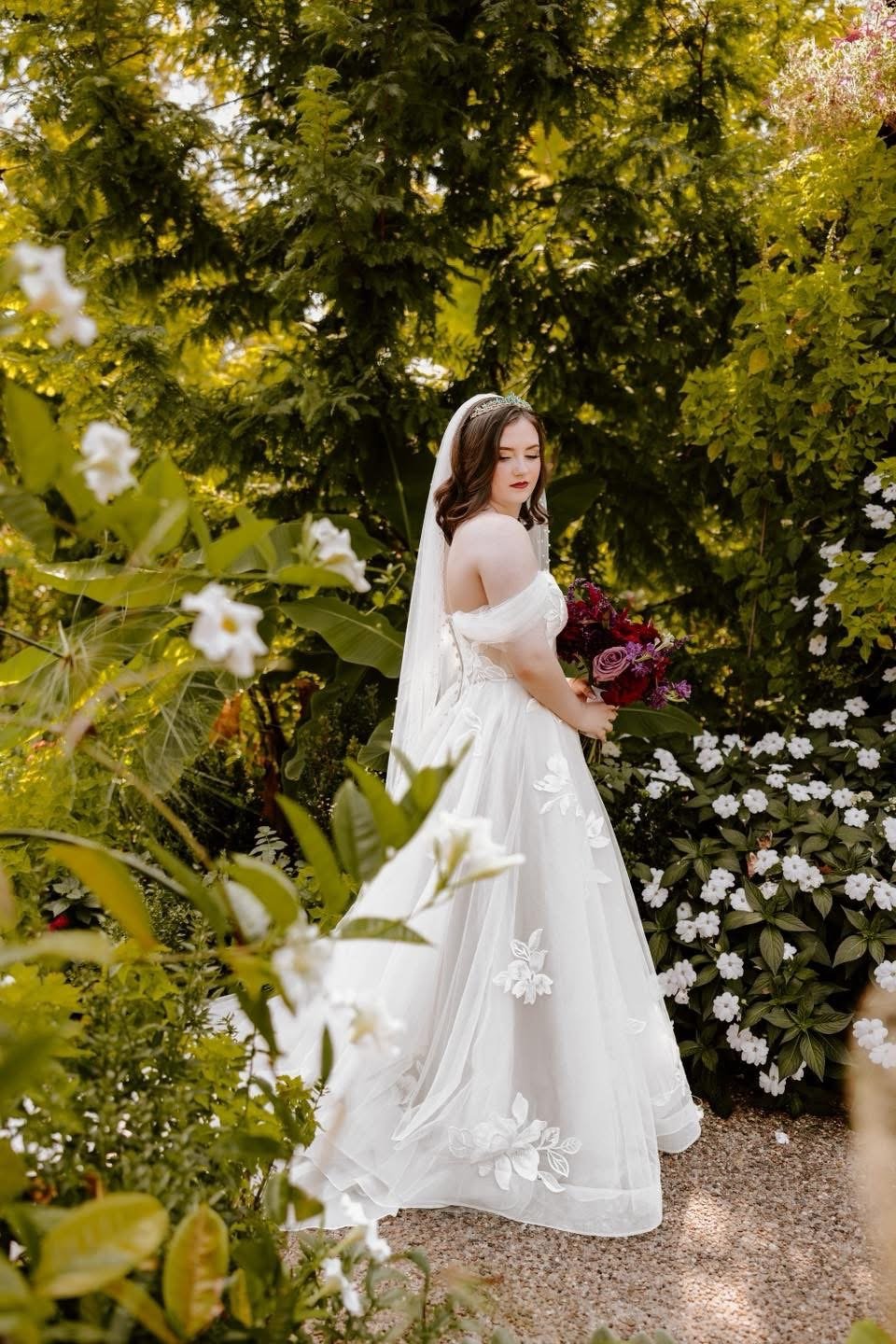 A bride in a white wedding gown holding a bouquet of dark red and purple flowers, standing outdoors amidst green foliage and white blossoms.