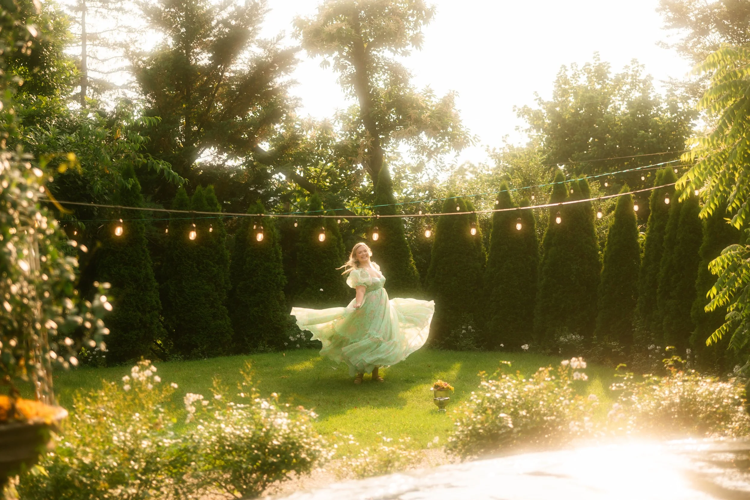 A woman in a flowing pastel dress spinning in a garden with string lights, surrounded by tall evergreen trees and flowering bushes, sunlight shining through.