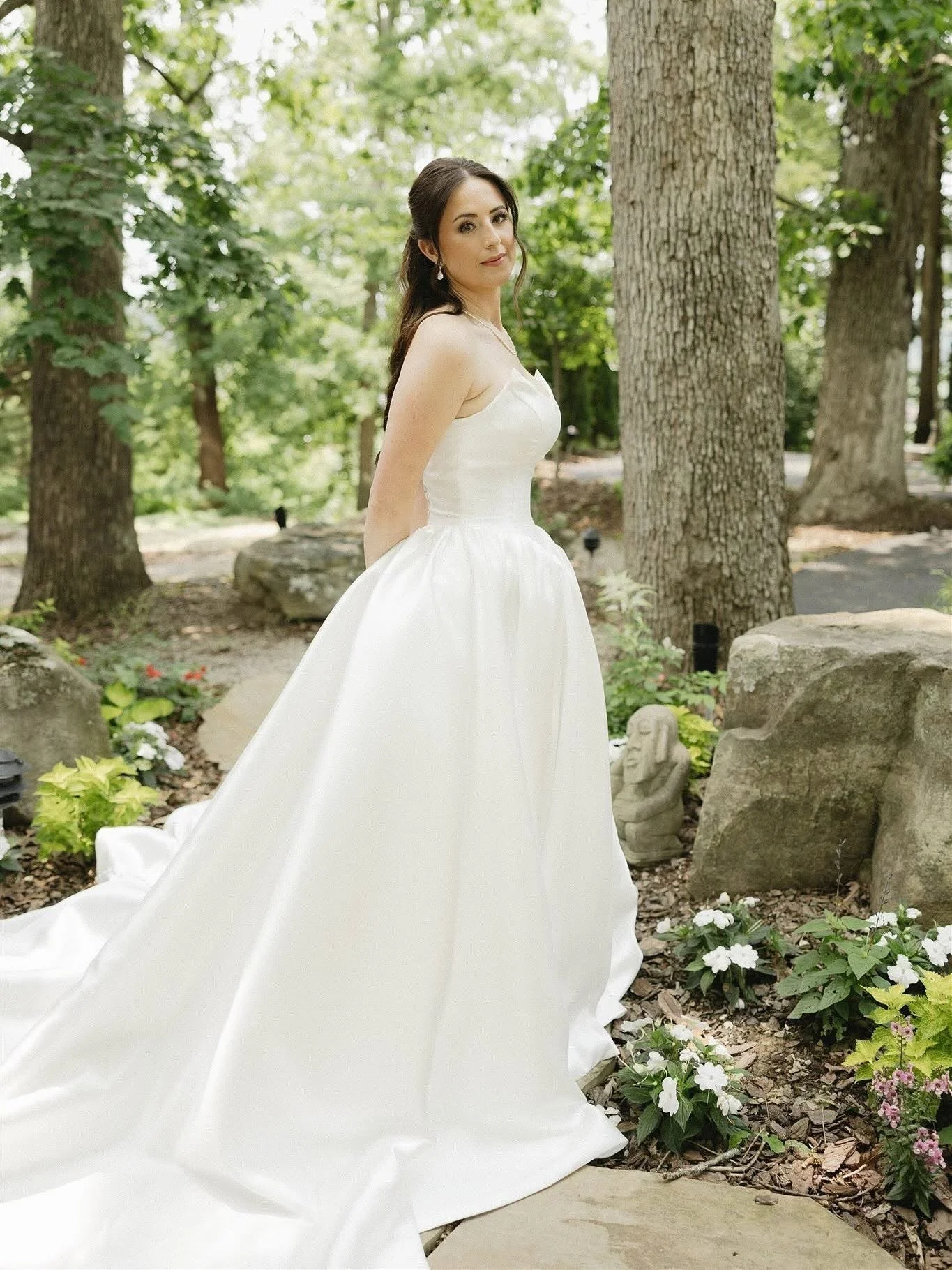 A woman in a white wedding dress standing outdoors among trees and rocks, with flowers and garden decor around her.